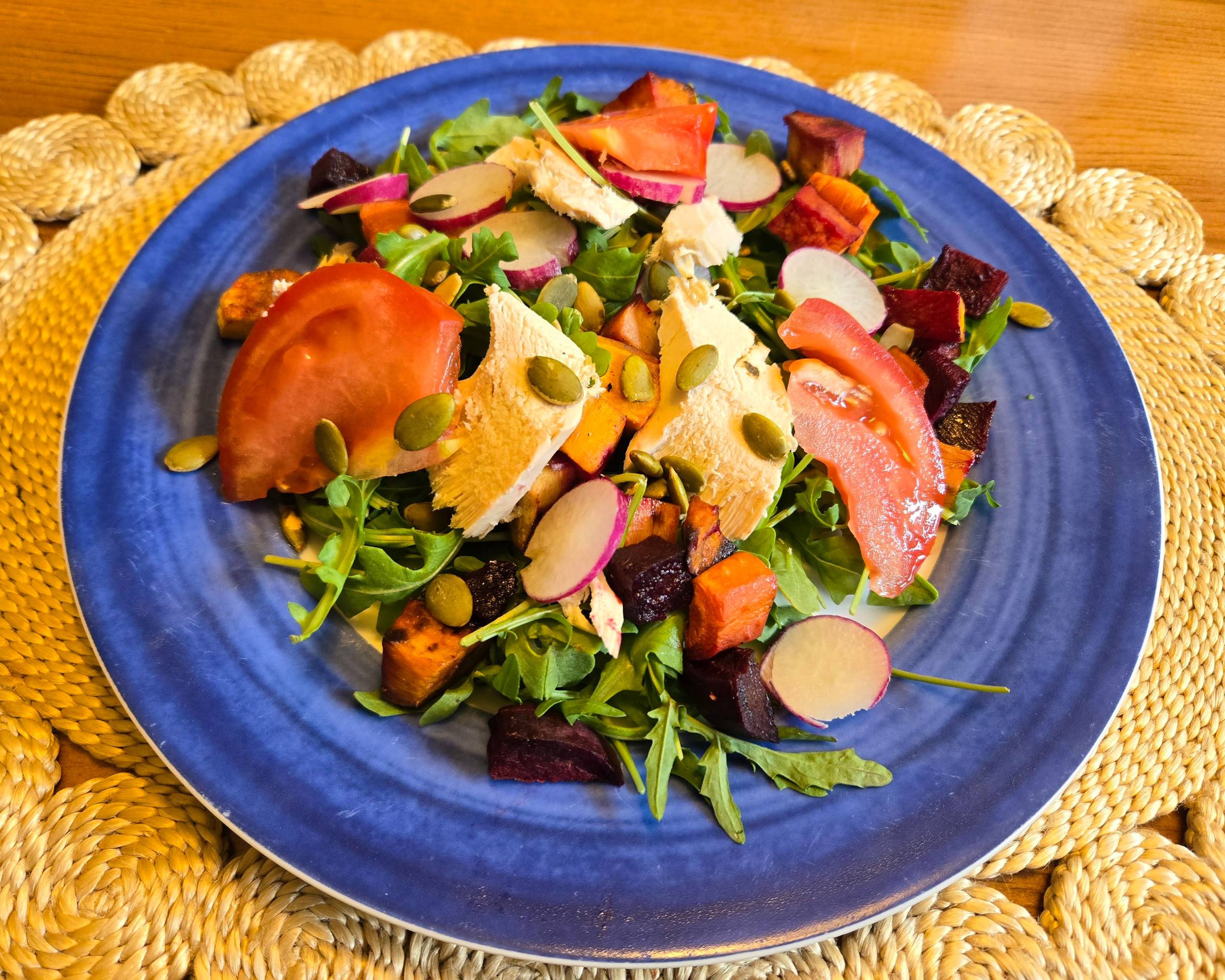 Salad with chicken, tomatoes, radishes, sweet potatoes, beets, greens, and pumpkin seeds on a blue plate.