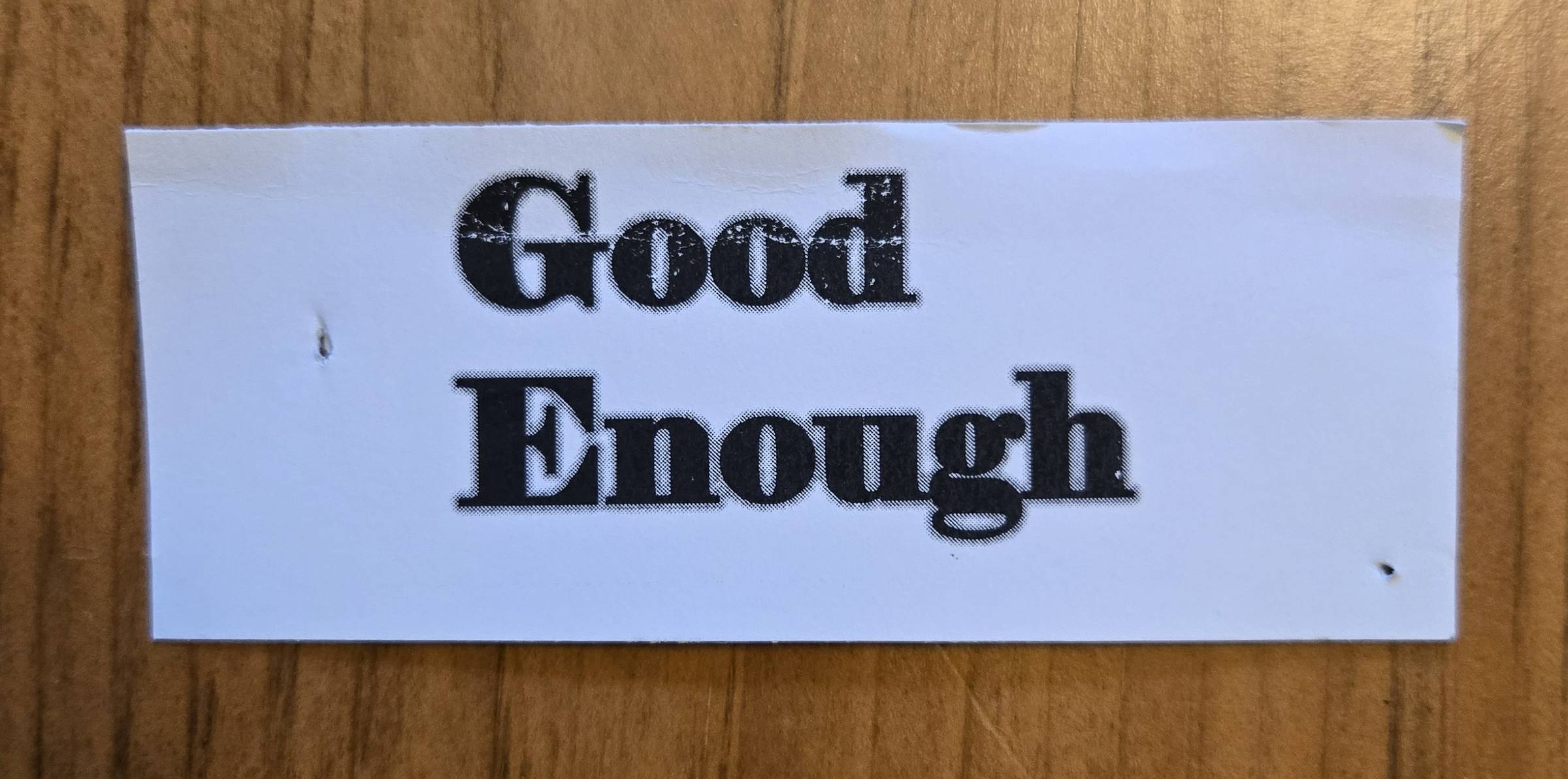 A white paper with "Good Enough" printed in bold black letters, placed on a wooden surface.