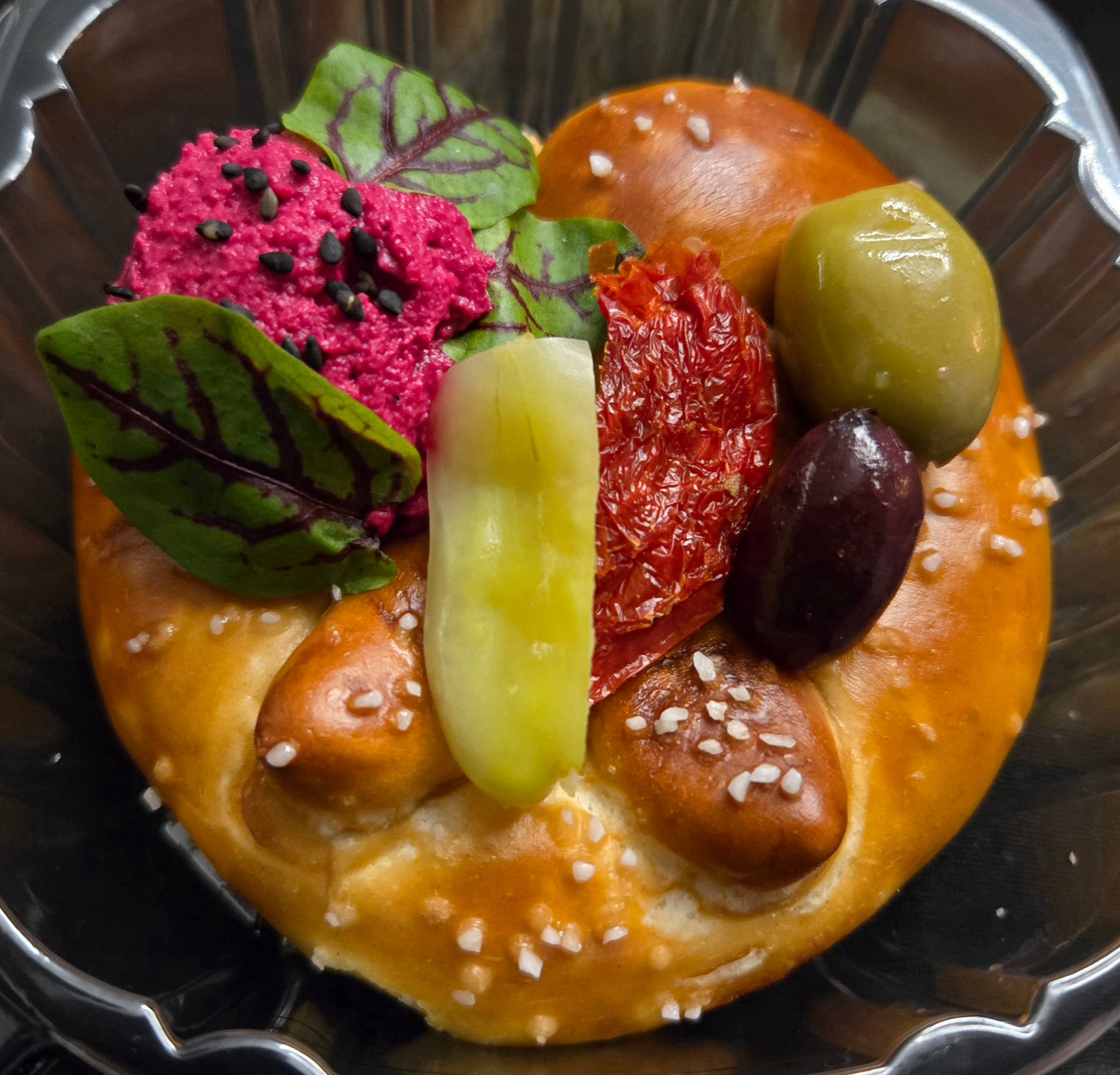 Gourmet pretzel topped with beet dip, olives, pickled vegetables, and herbs in a small black dish.