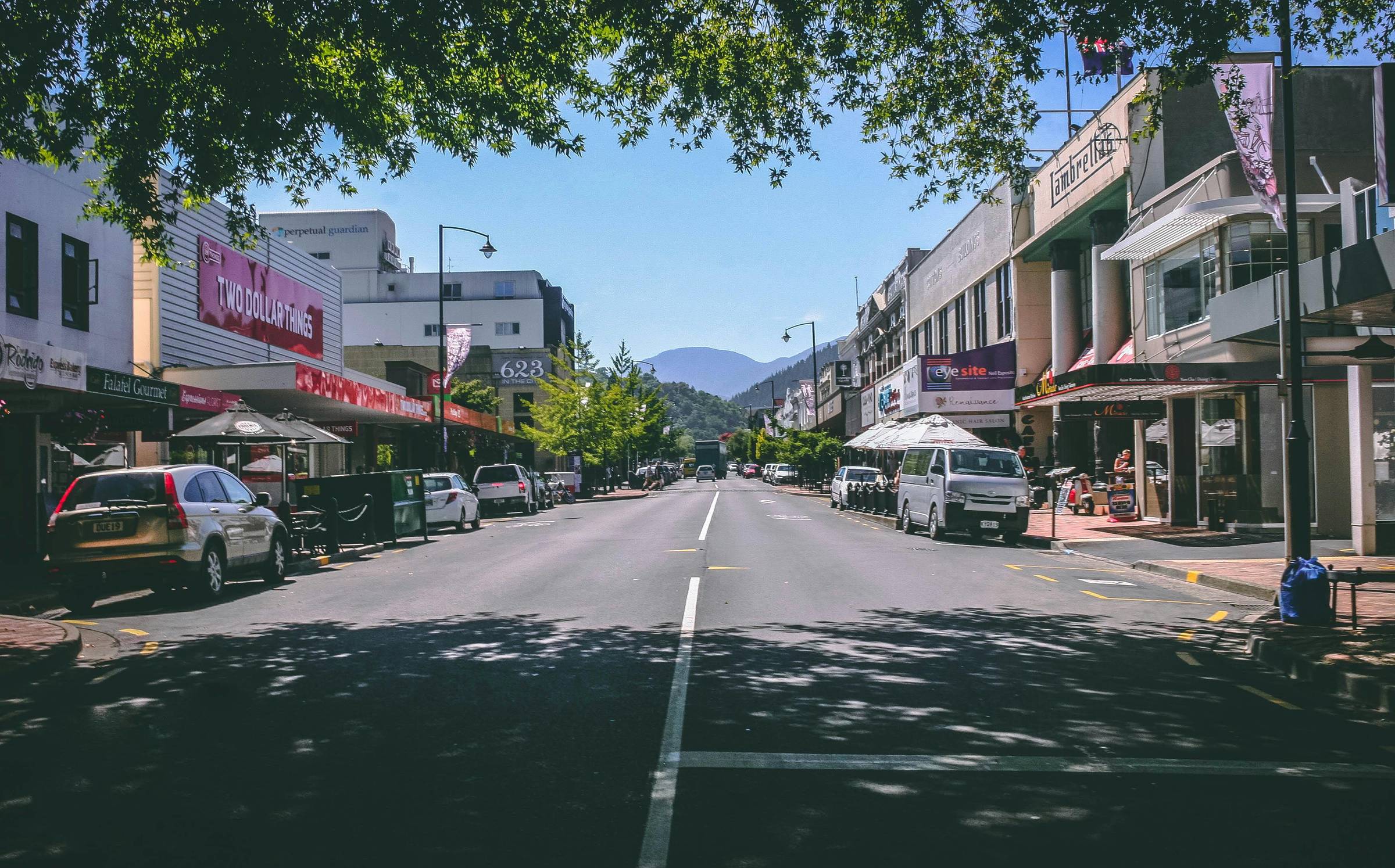A quiet city street with parked cars, shops, distant mountains, and some shade from trees.