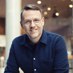 A person wearing glasses and a blue shirt is smiling, sitting indoors with a blurred background featuring warm lighting.