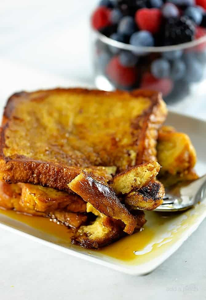 Stack of French Toast with maple syrup on a white platee with mixed berries in a glass bowl in the background.