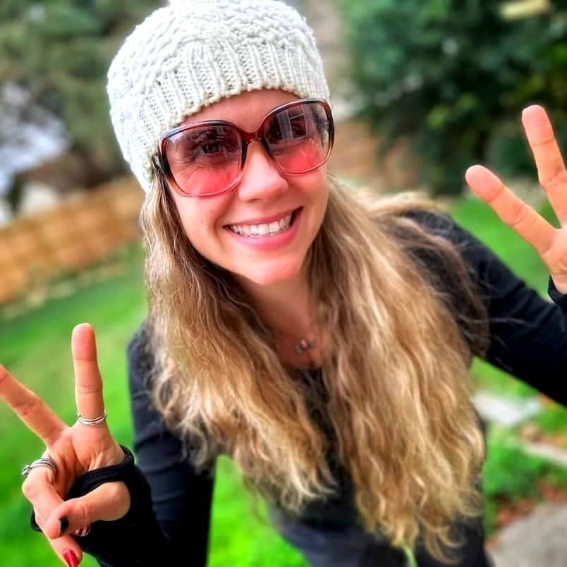 A person with long, wavy hair is smiling and wearing a white knit beanie and sunglasses. They are dressed in a black top and making peace signs with both hands. The background is an outdoor setting with green grass and blurred trees.