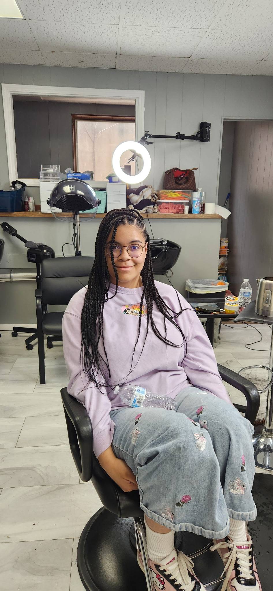 A young teen with brown skin and long black braids, sitting in a chair in a beauty shop. She is wearing a purple sweatshirt, blue jeans with bunnies and flowers on them and Hello Kitty sneakers.