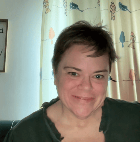 A picture of an older woman in her fifties with short hair and a green shirt, sitting in front of curtains with birds on them.
