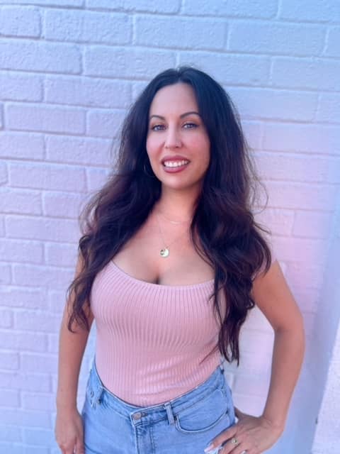 A white woman with long dark hair, wearing a pink top and a necklace.