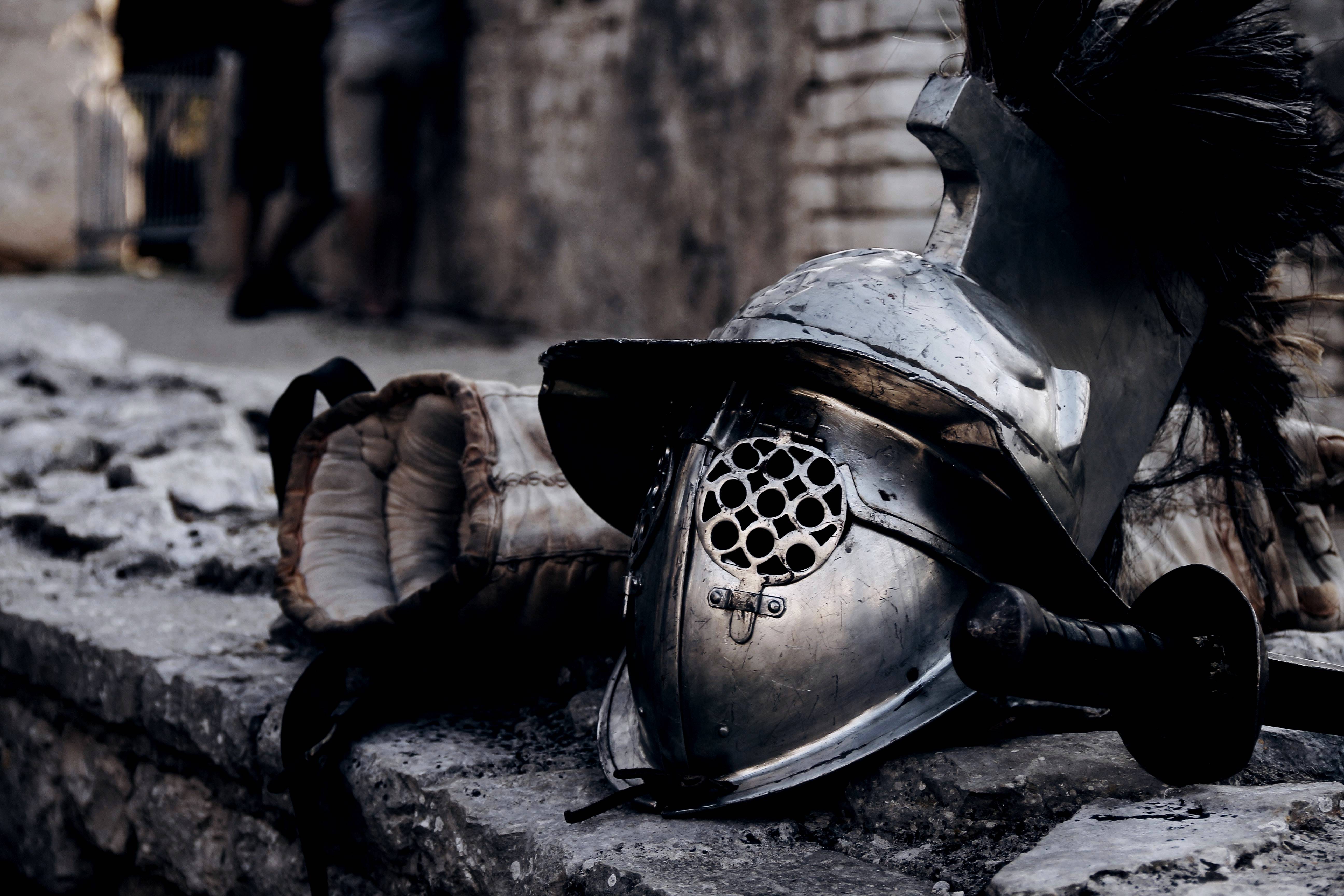 A metal gladiator helmet with a grill faceplate is resting on a stone surface, along with a pair of padded gloves and a short sword with a dark hilt. The helmet has a prominent crest, and the setting appears to be an ancient or historical location with stone architecture in the background.