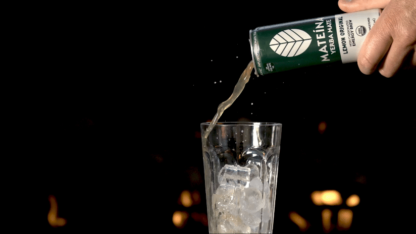 A hand pours a can of Mateina Lemon Original yerba mate into a glass filled with ice cubes against a dark background with blurred lights.