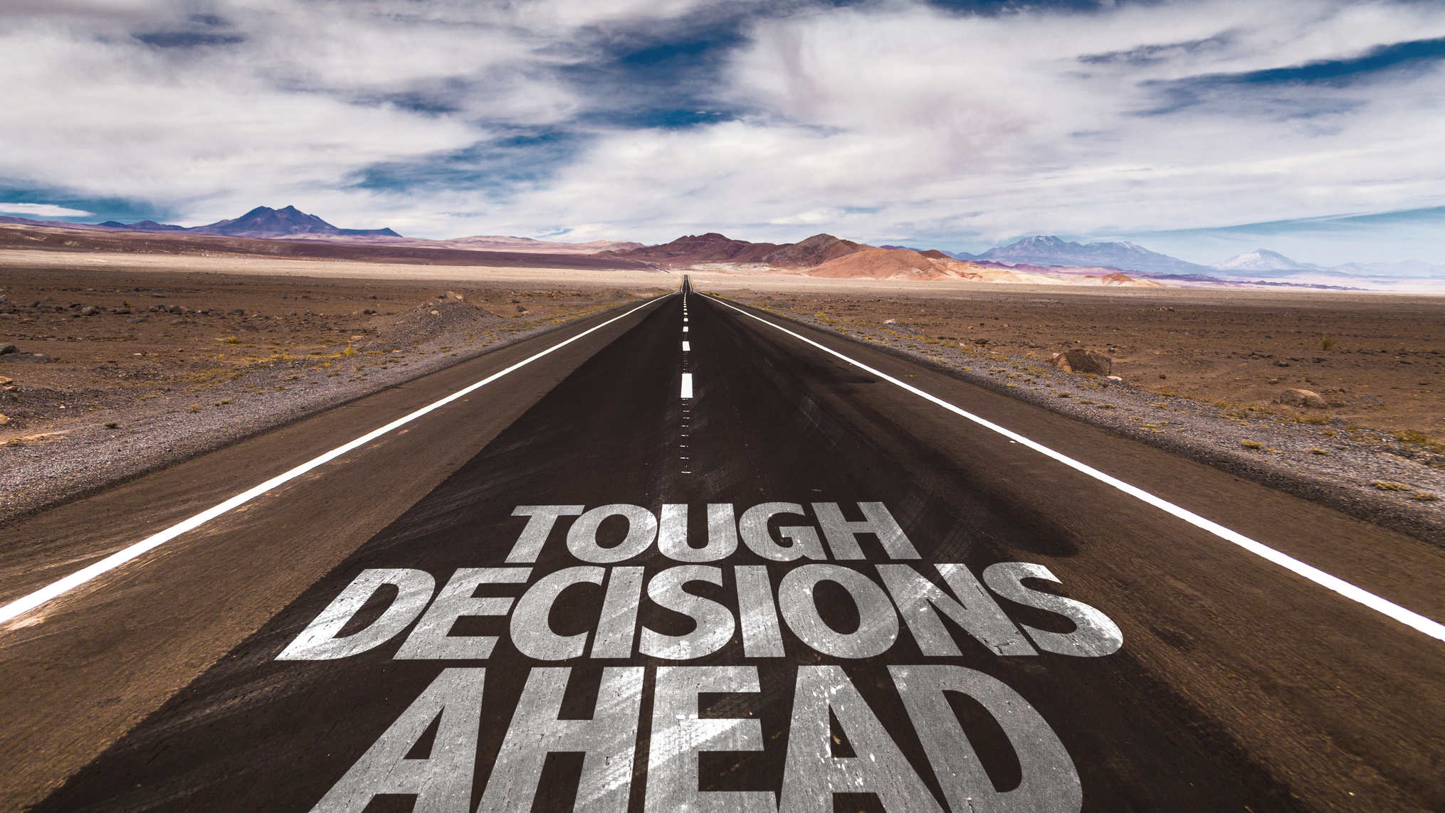 A straight two lane road cuts through a desert landscape toward distant mountains under a wide sky of broken clouds, with the words TOUGH DECISIONS AHEAD painted in large white block letters across the asphalt in the foreground. The concept is the weight of leadership choices, showing that the road forward is clear and open but demands hard calls from whoever is driving.