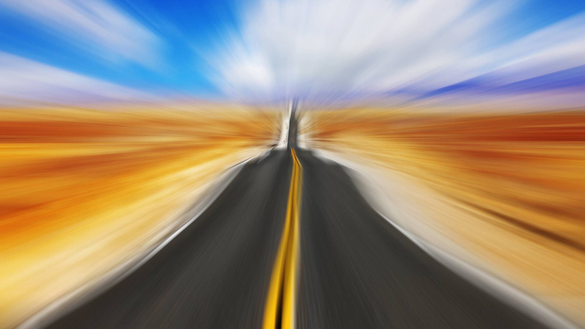 A long open highway with a vivid yellow centre line stretches toward a vanishing point on the horizon, captured with intense radial motion blur that pulls the eye forward. Golden fields blur outward on either side of the road, while a brilliant blue sky with streaks of white cloud opens above. The concept captures the velocity at which leadership decisions are made, illustrating how speed without clarity can carry an organisation forward without ever asking whether it is moving in the right direction.