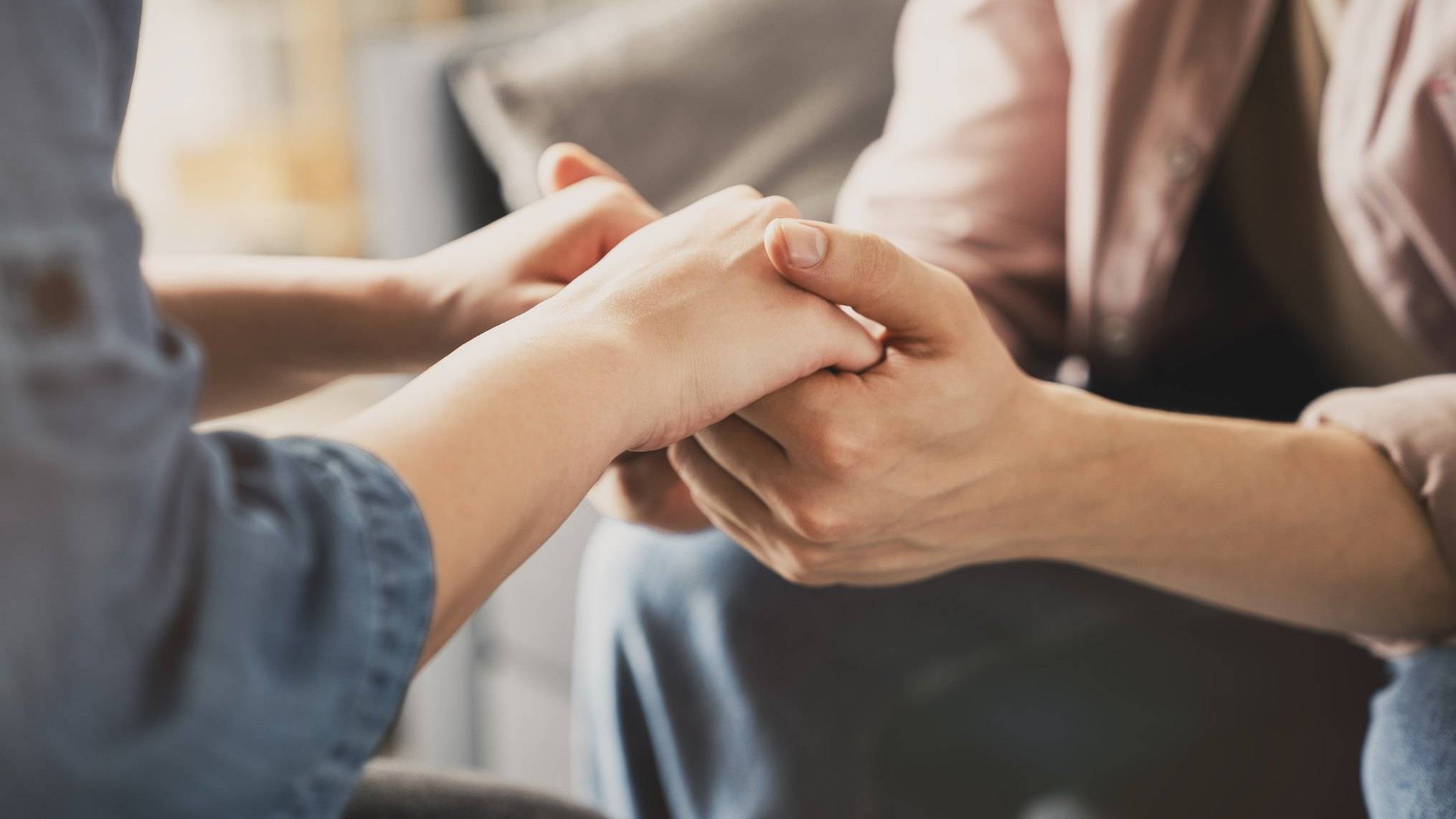Close up of two people sitting together as one person holds the other's hand in both of theirs, with soft window light behind them. The image represents care that shows up as presence and attention rather than performance.
