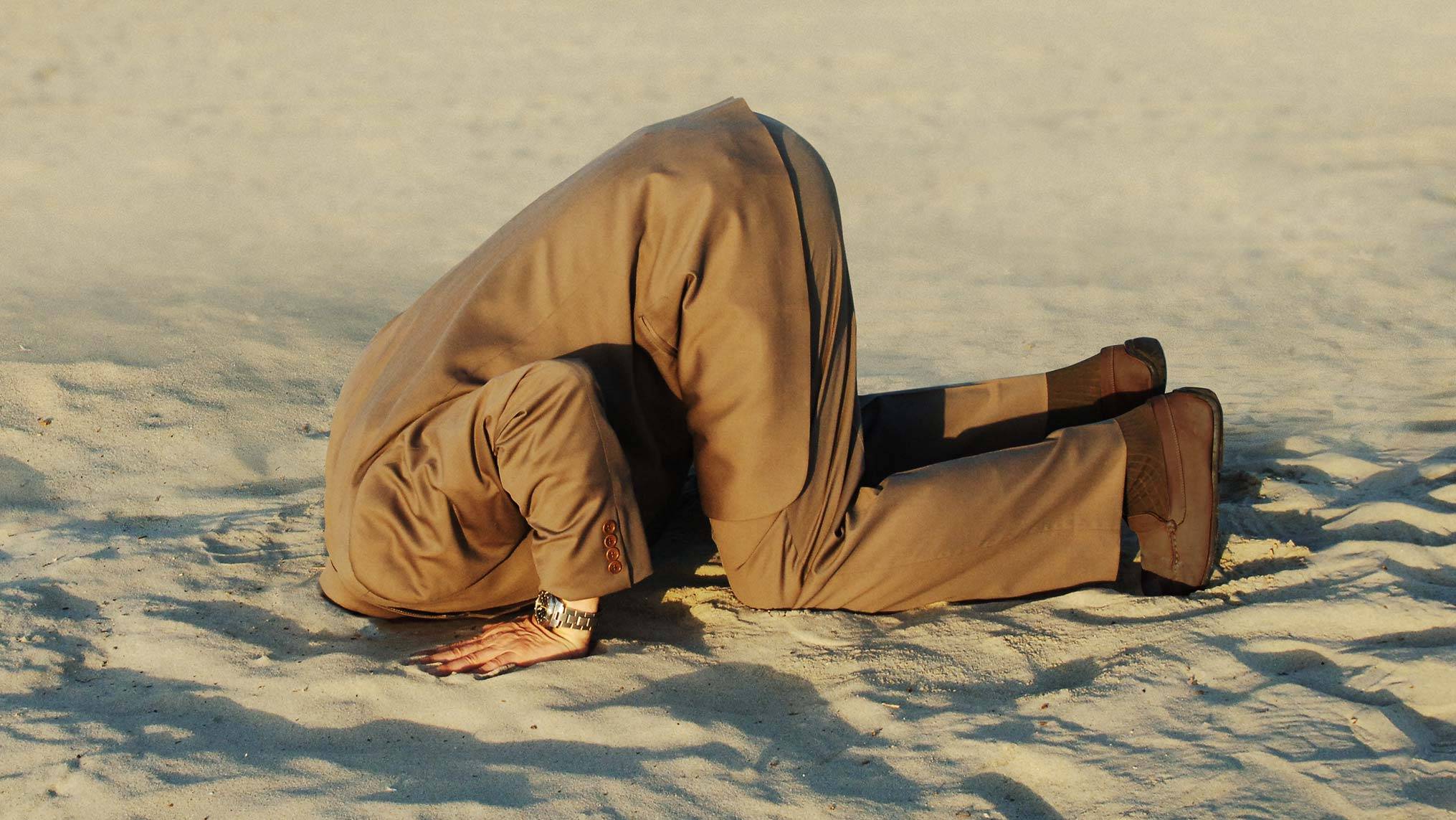 Man in business suit burying his head in the sand, illustrating leadership avoidance and the refusal to face difficult realities.
