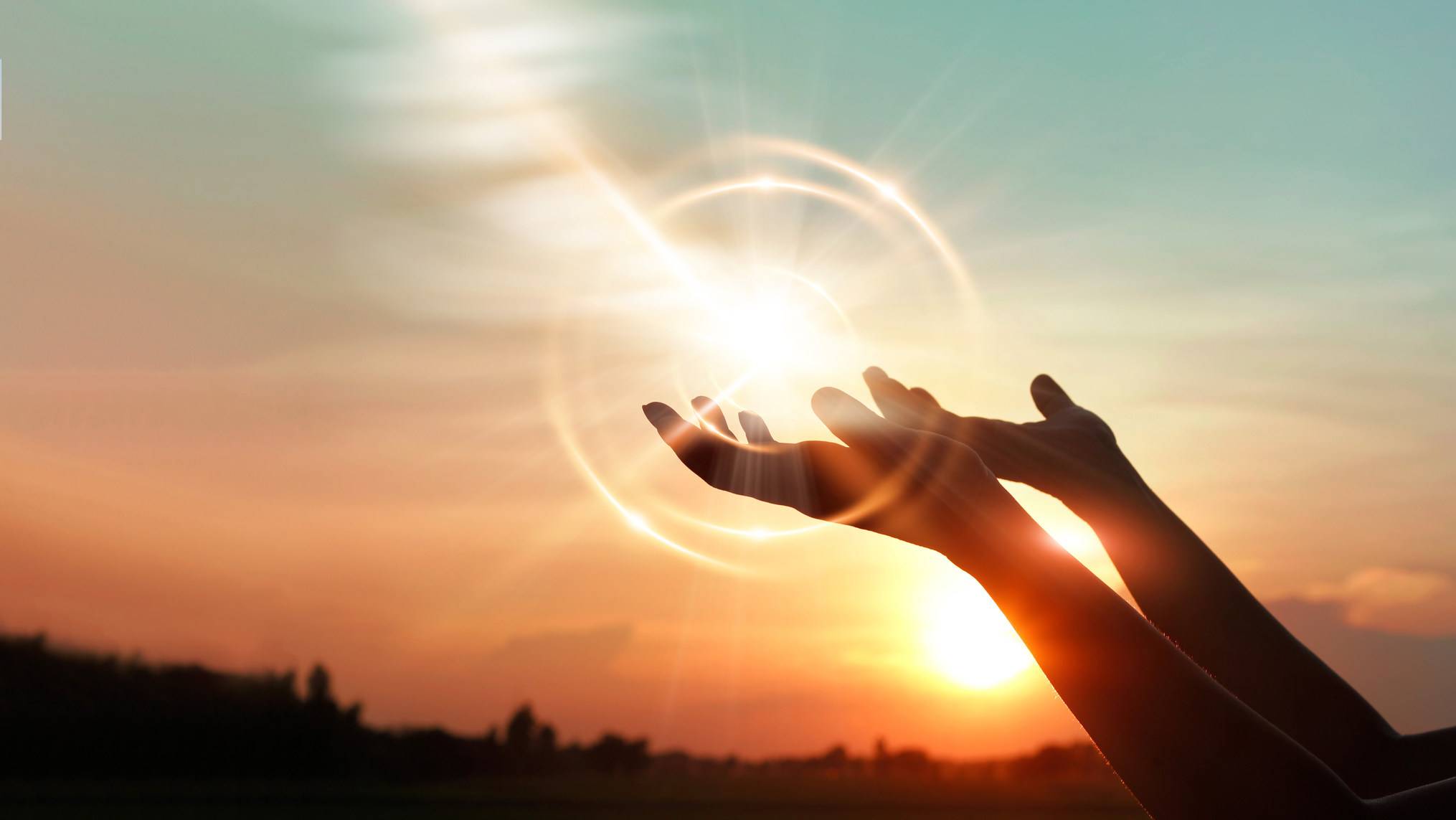 Two open hands reach upward toward a glowing sun surrounded by rings of light against a warm sunset sky over a dark treeline. The image represents openness, hope, and reaching toward something larger than yourself.
