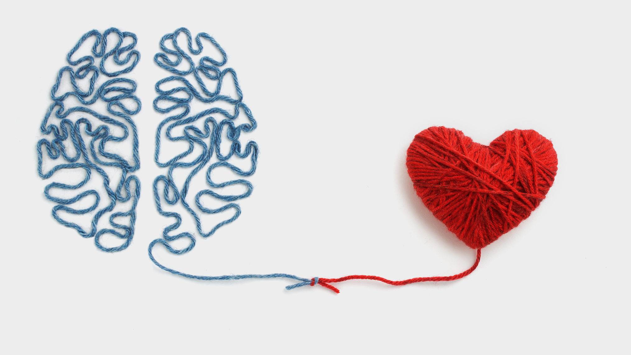A brain made from looped blue yarn sits on a white background beside a heart wound from red yarn, with a single strand from each reaching across the empty space to meet and tie together in a small knot. The concept is the connection between thinking and feeling, showing that emotional intelligence is the thread that links reason and emotion rather than choosing one over the other.