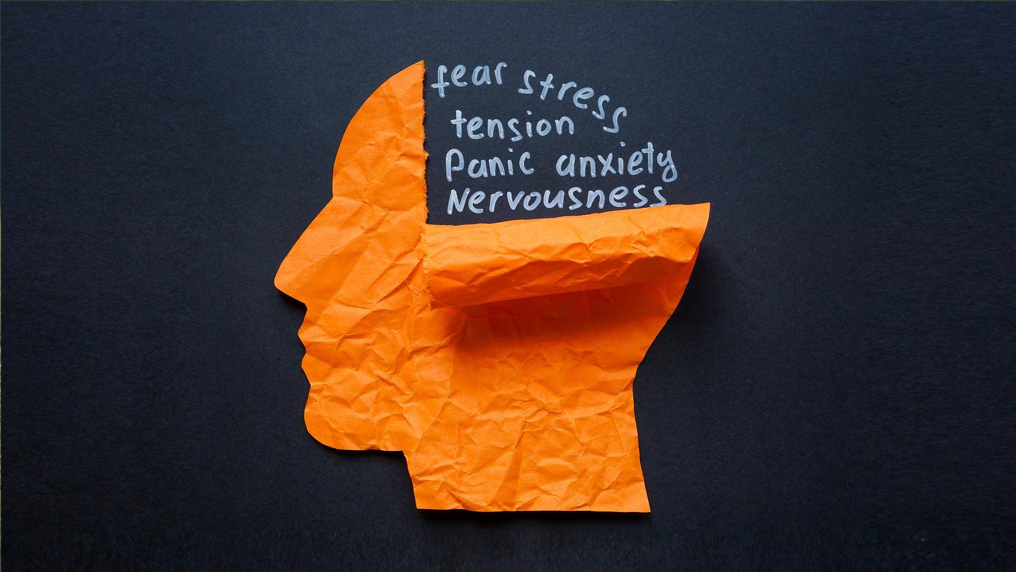 A crumpled orange paper cutout shaped like a human head in profile rests against a dark charcoal background, with the top of the head torn open to expose the dark space inside. Written in white chalk style handwriting within the opening are the words "fear, stress, tension, panic, anxiety, nervousness." The concept represents the internal weight of unprocessed emotions, illustrating how anxiety and stress crowd the mind when there is no space to slow down and respond rather than react.