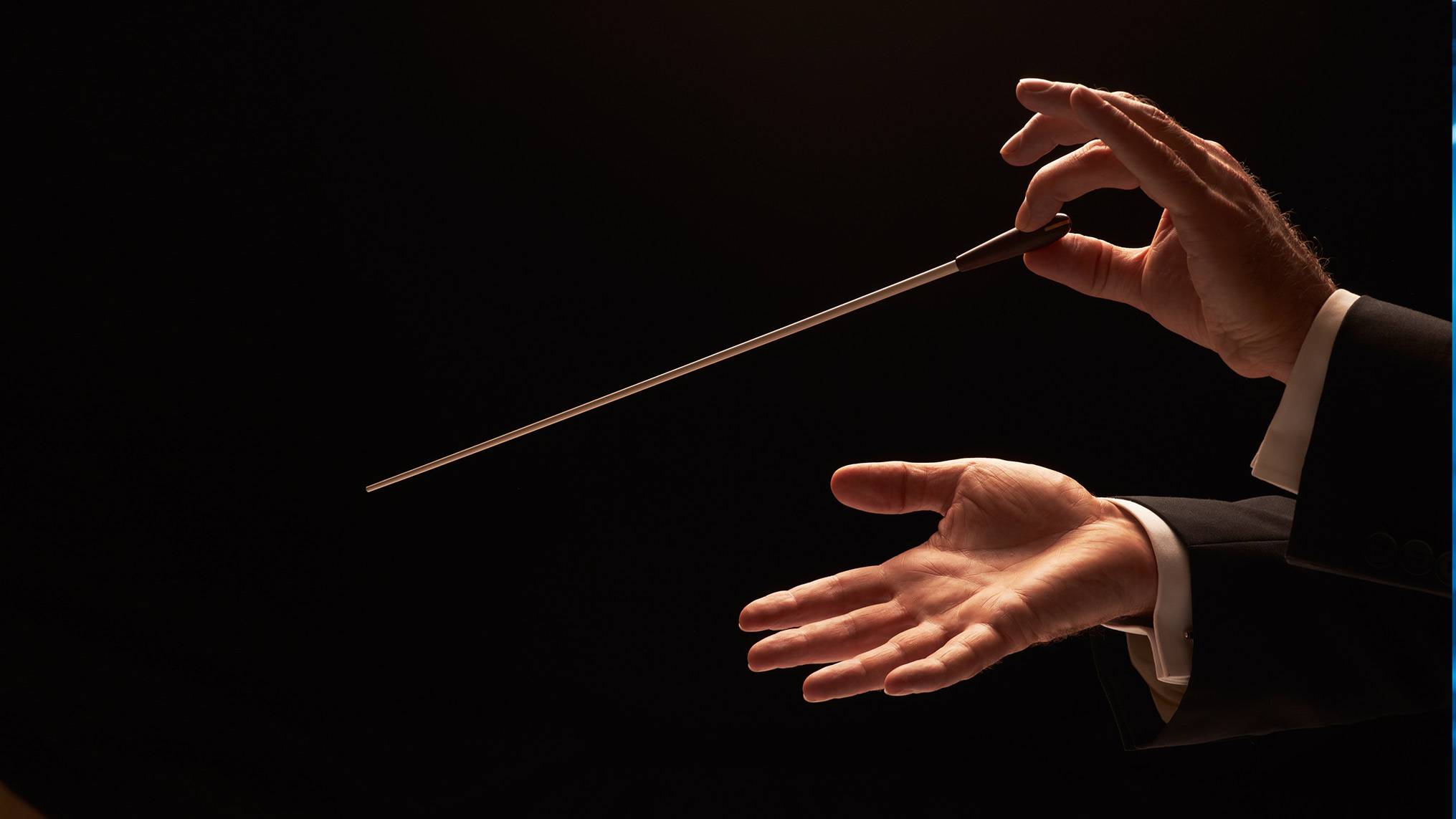 A pair of weathered hands emerge from the darkness wearing the black sleeves and white cuffs of a formal tuxedo, with one hand holding a conductor's baton and the other open and expressive against a pitch black background. The image represents the art of directing without dominating, illustrating the concept that true leadership is about guiding the rhythm, unity, and performance of others rather than playing every note yourself.