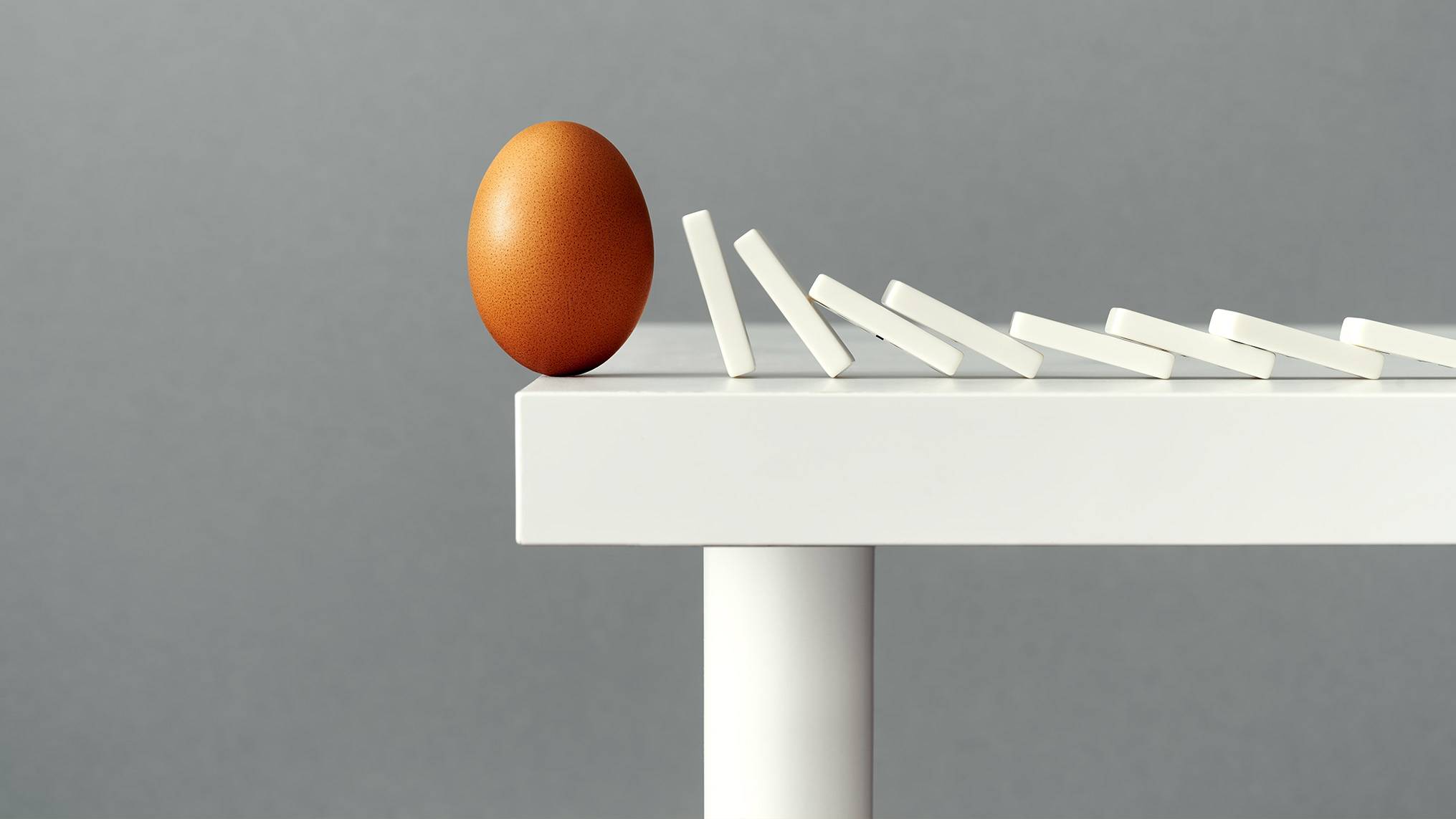 A single brown speckled egg stands upright on the edge of a white table next to a line of white dominoes that are falling in sequence away from it, set against a soft gray background. The image represents how one small, fragile element can trigger a chain reaction, illustrating the concept that emotional intelligence begins with recognizing the delicate moments that set larger consequences in motion.