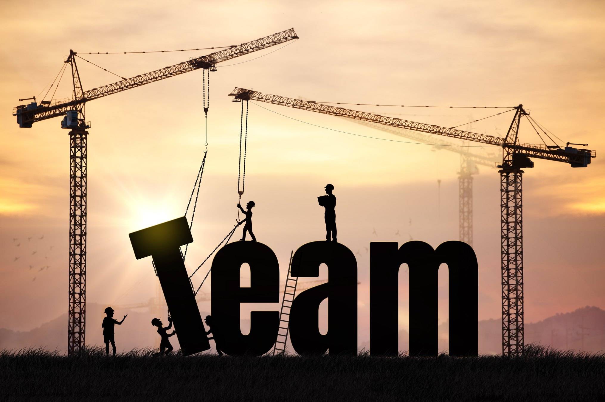 Silhouettes of construction workers and two tall cranes assemble giant black letters spelling the word Team against a golden sunset sky, one worker guiding the letter T into place while others stand on top of the letters. The image represents culture as something leaders build through repeated action rather than declare on a slide.