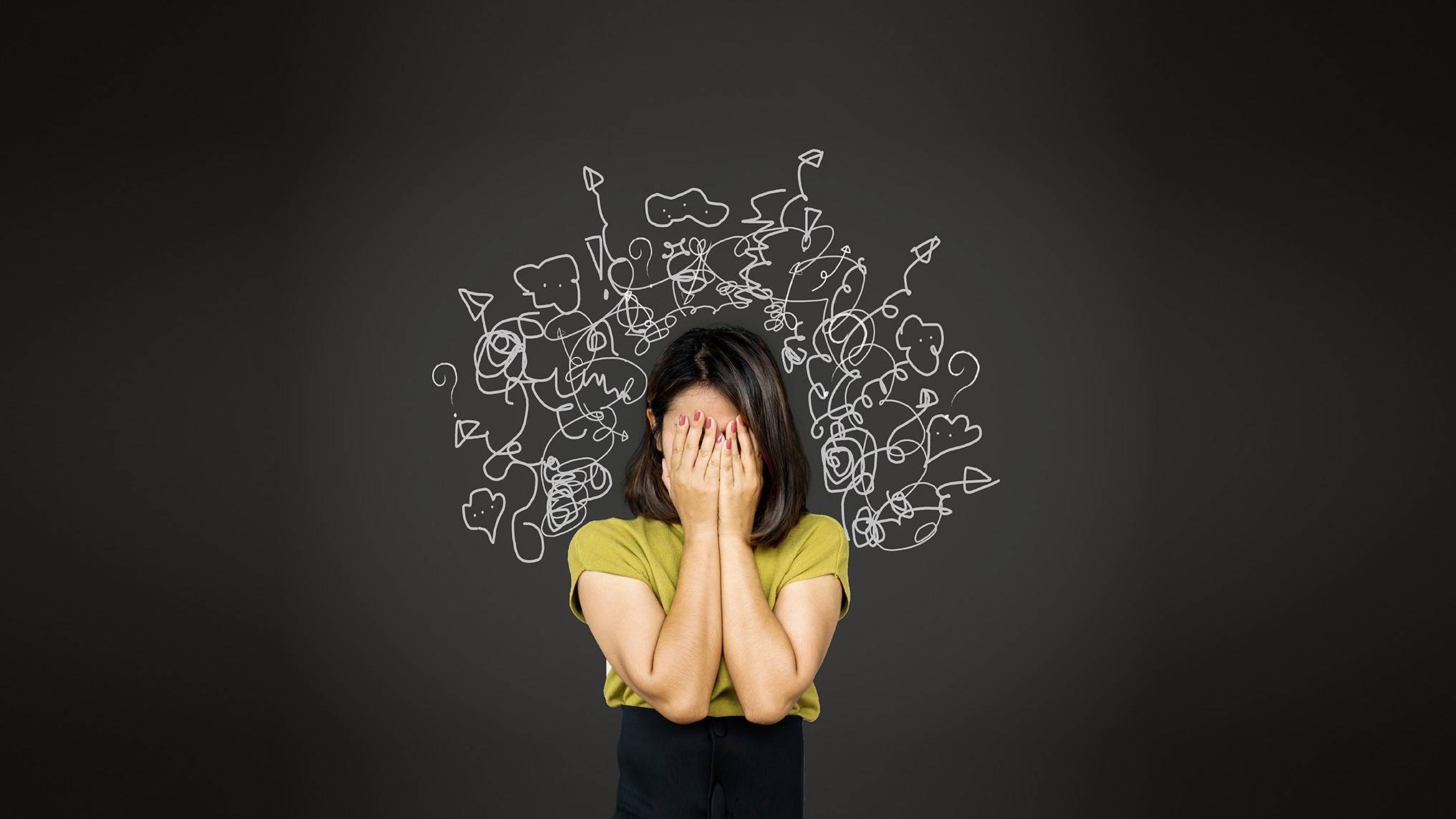 A woman in a mustard yellow short sleeve top with shoulder length dark hair stands against a dark charcoal gray background, covering her face with both hands. White hand drawn doodles, tangled scribbles, question marks, exclamation points, and chaotic squiggles swirl outward from behind her head like a cloud of mental noise. Mental overwhelm and emotional flooding. The image visualizes the internal chaos of a mind overrun by unprocessed thoughts, stress, and competing demands, illustrating what it feels like when emotional load exceeds capacity.