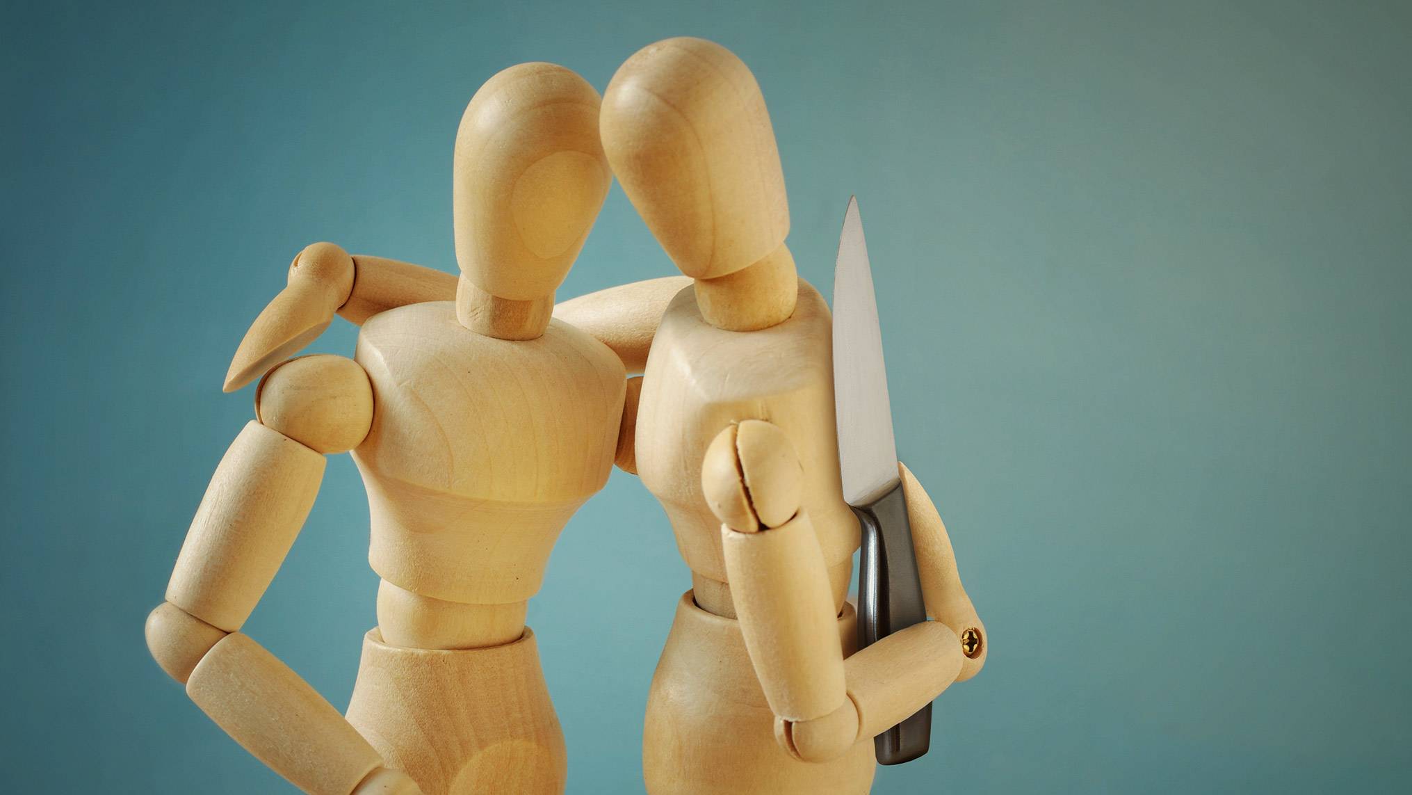 Two wooden artist mannequins embracing, with one holding a knife behind the other's back, set against a teal background. The embrace represents the appearance of closeness and trust, while the hidden knife represents the subtle betrayals happening beneath the surface. The danger is invisible to the person being held, mirroring how micro-betrayals operate undetected within relationships that still look intact.