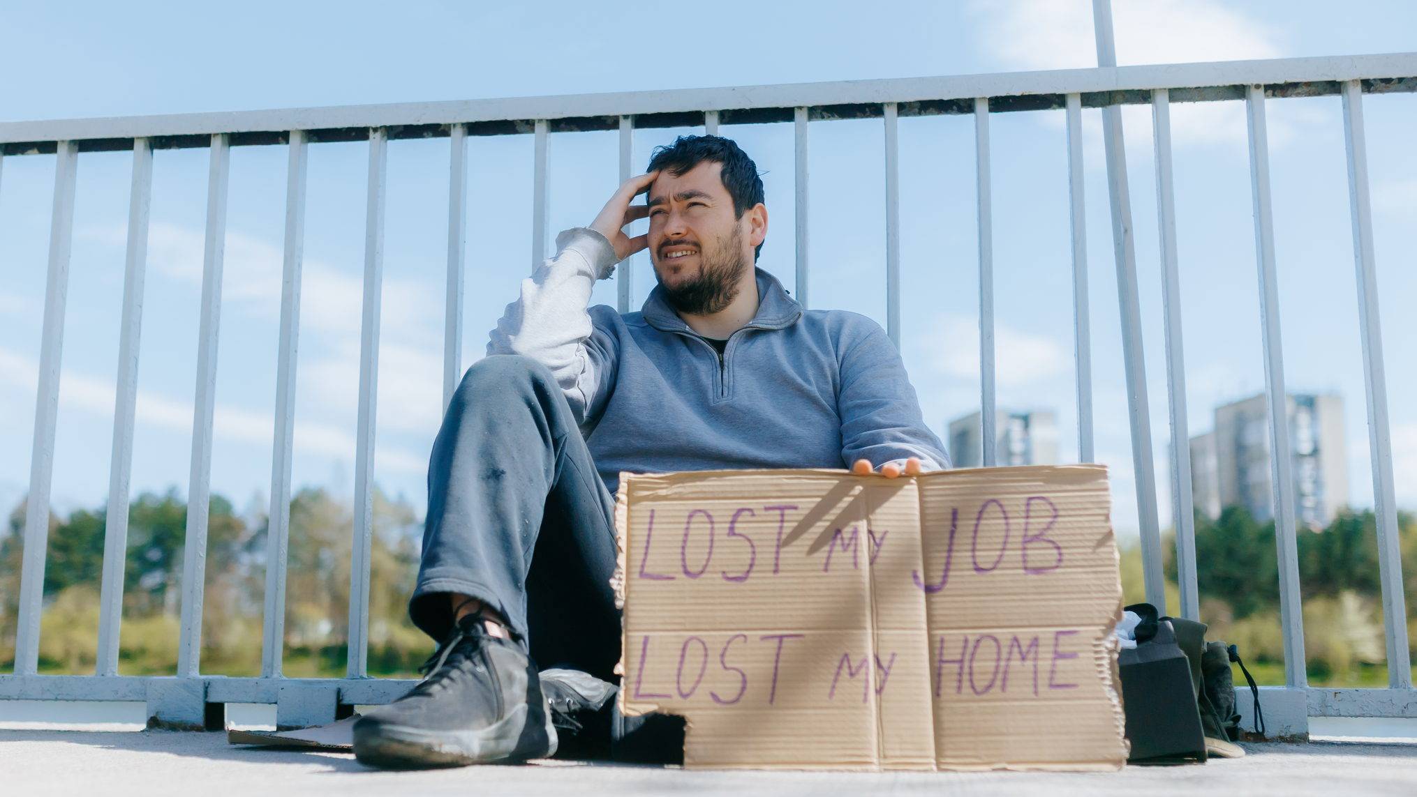 Visual description: A bearded man in a grey sweatshirt sits on concrete against a white railing under a bright blue sky, one hand pressed to his forehead, a hand lettered cardboard sign propped in front of him reading LOST MY JOB LOST MY HOME, with a backpack beside him and city towers in the distance. Concept: The hardest chapters arrive without warning and without dignity, and they look nothing like the polished growth stories we are sold. Development happens inside the wreckage, not after it has been cleaned up.