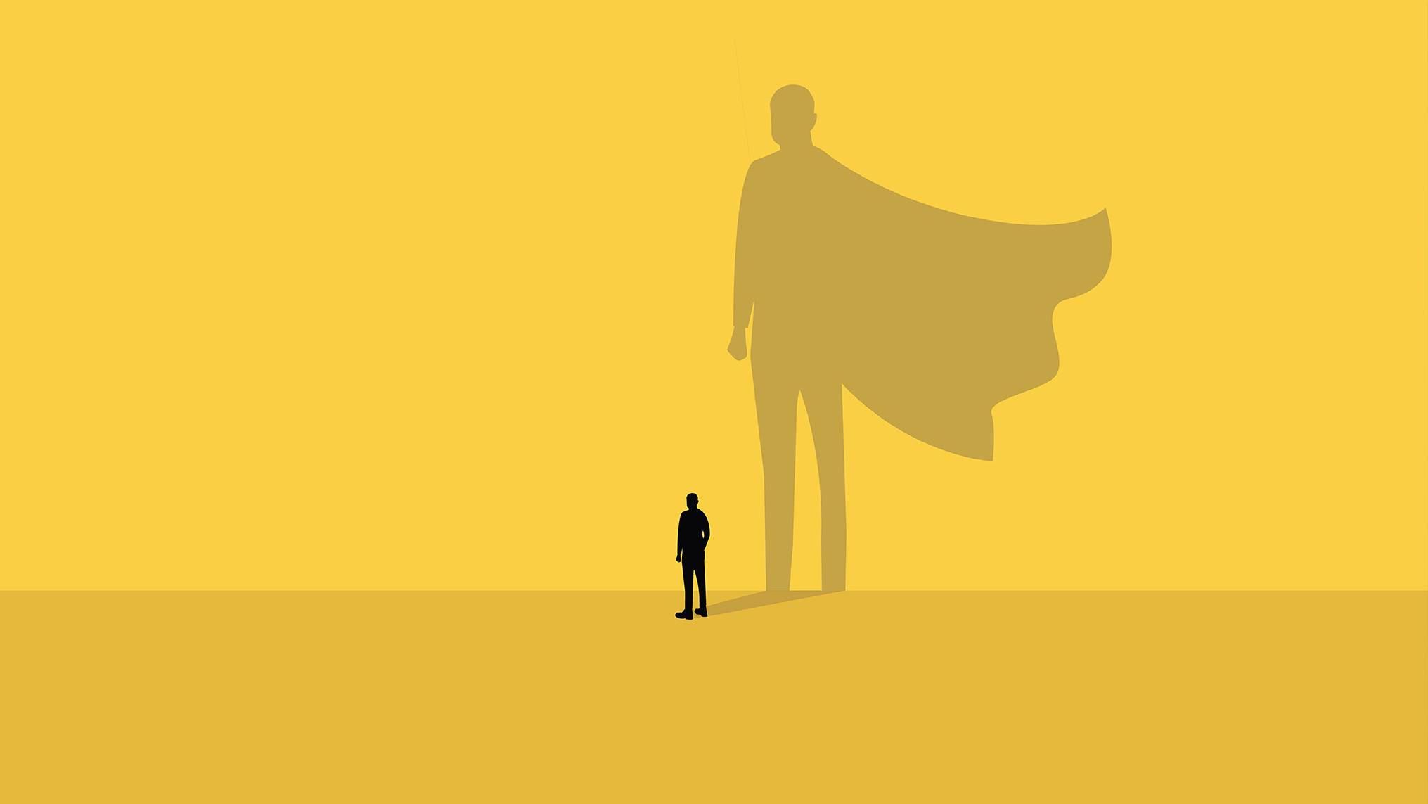 A small silhouetted figure of a man stands on a yellow background while casting a towering shadow of himself wearing a flowing superhero cape. The image represents the gap between how we see ourselves in the present and the greater potential that lives within us, illustrating the concept that personal development is the journey of growing into the larger version of who we are capable of becoming.