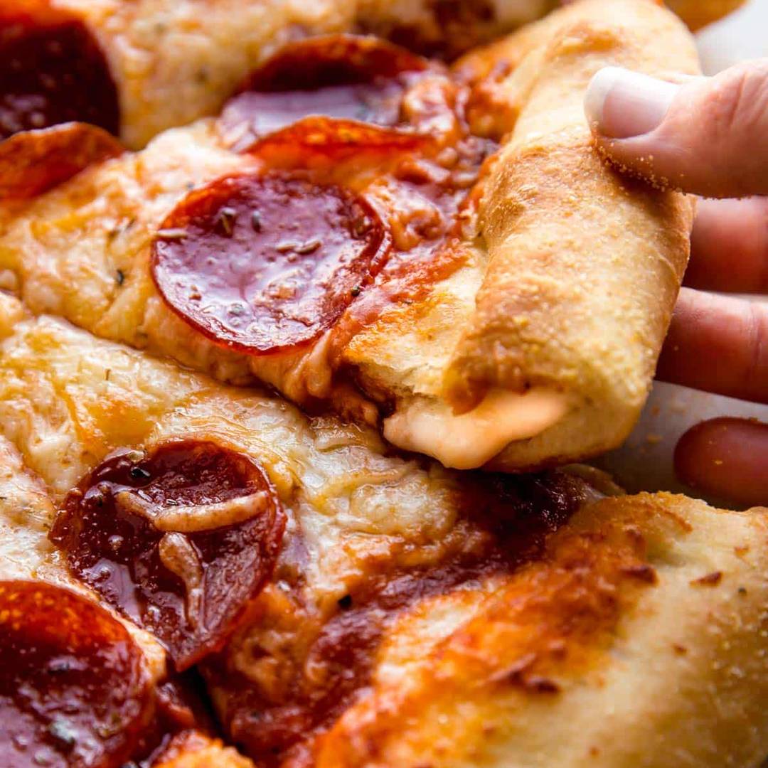 a hand pulling up a piece of stuffed crust pizza with pepperoni
