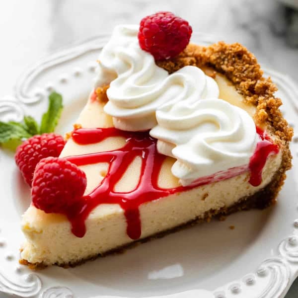 slice of cheesecake pie on a plate topped with whipped cream, raspberries, and raspberry sauce