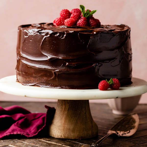 a layer cake on a cake stand, covered in chocolate ganache and topped with fresh raspberries