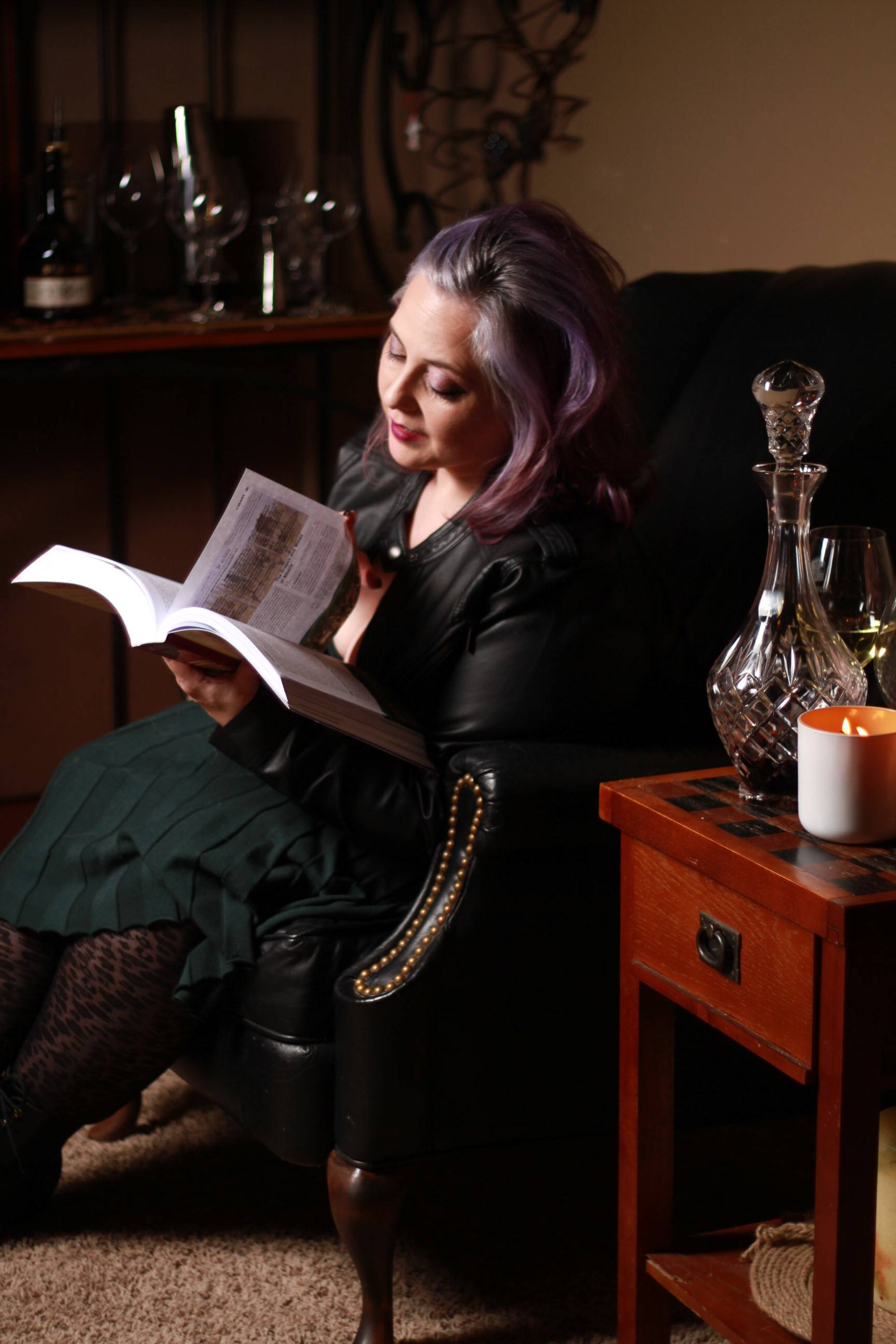 Woman with lavender hair sits in a black leather armchair reading a large astrology book, surrounded by cozy decor including a lit candle, wine glass, and crystal decanter on a wooden side table.