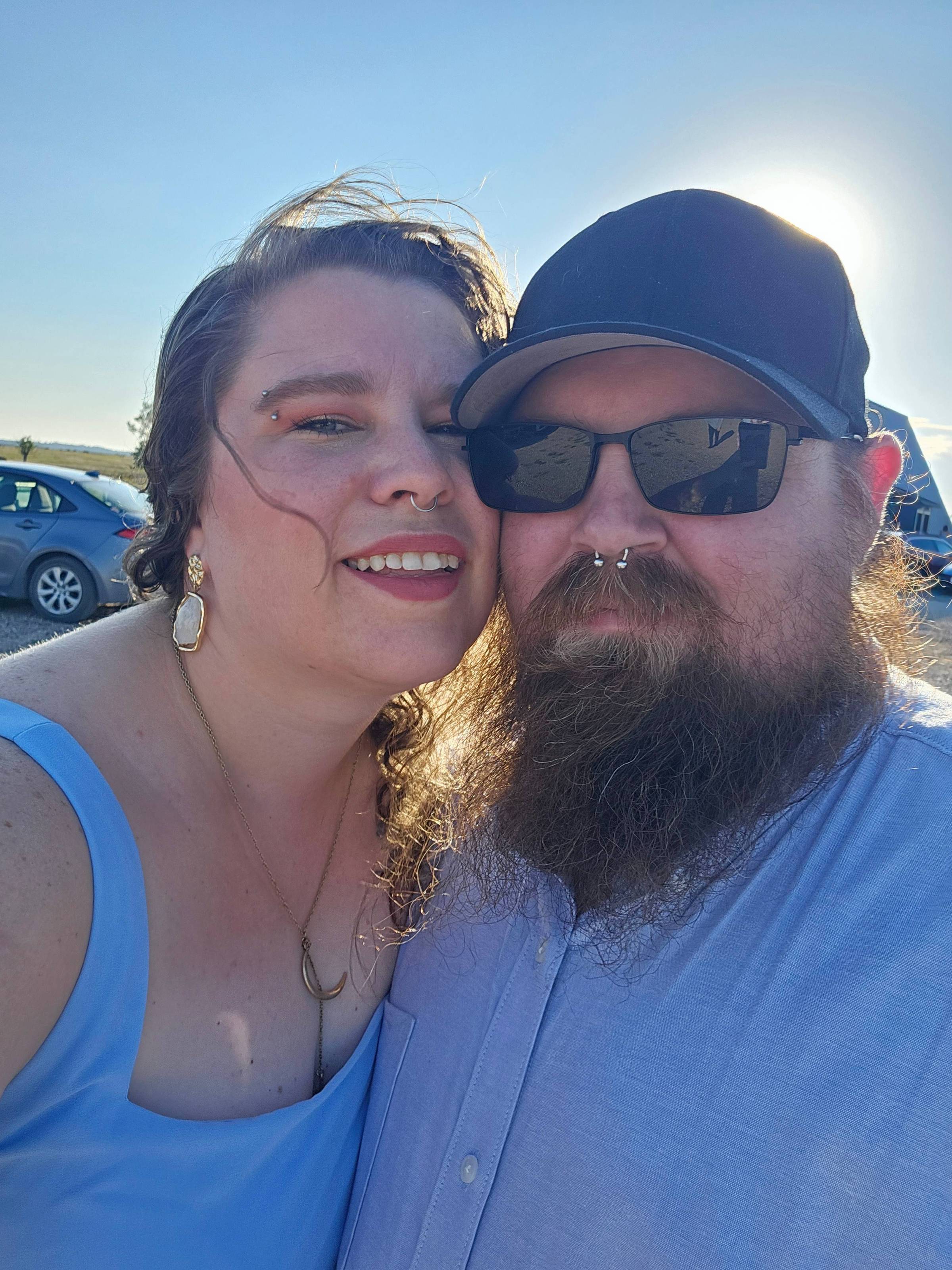 Shanae and Jeremy together outdoors on a sunny day at a wedding ceremony. She is wearing a light blue dress, gold jewelry, and has curly hair. He is wearing a light blue shirt, black cap, dark sunglasses, and has a full beard. The sun is shining behind th