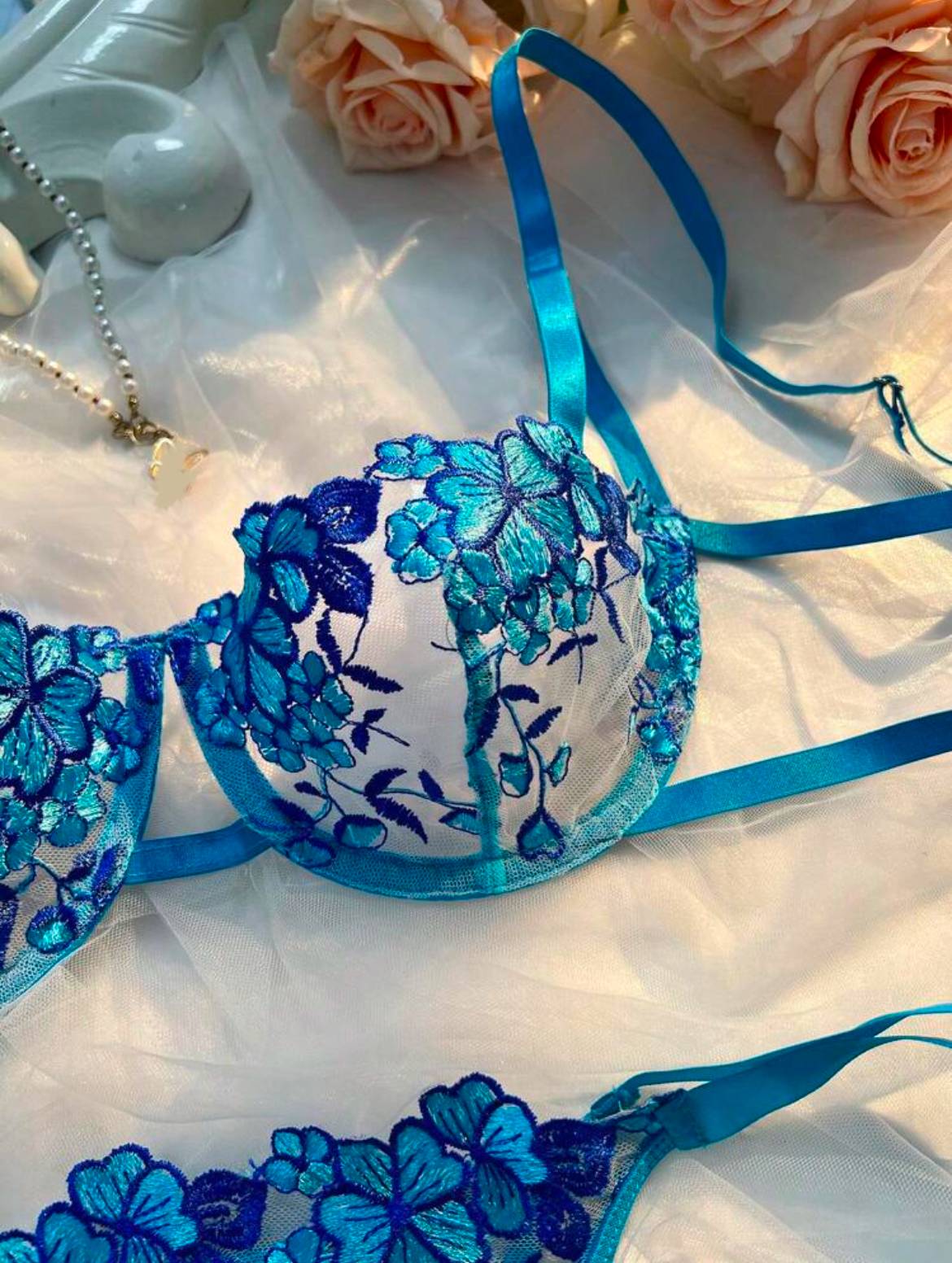 A blue lingerie set featuring floral embroidery is laid out on a light-colored, textured fabric. The set includes a bra and panties with sheer material and blue straps. A decorative piece is partially visible in the background.