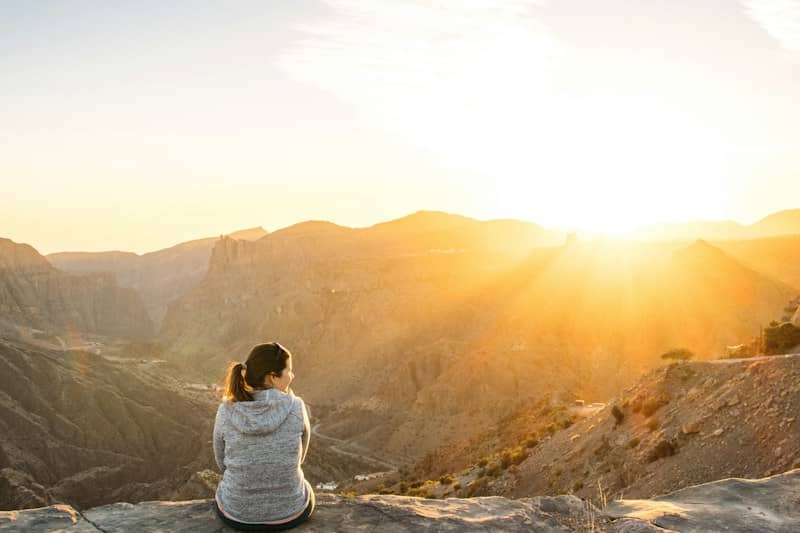 A person in a grey hoodie sitting on a rocky cliff overlooking mountains
