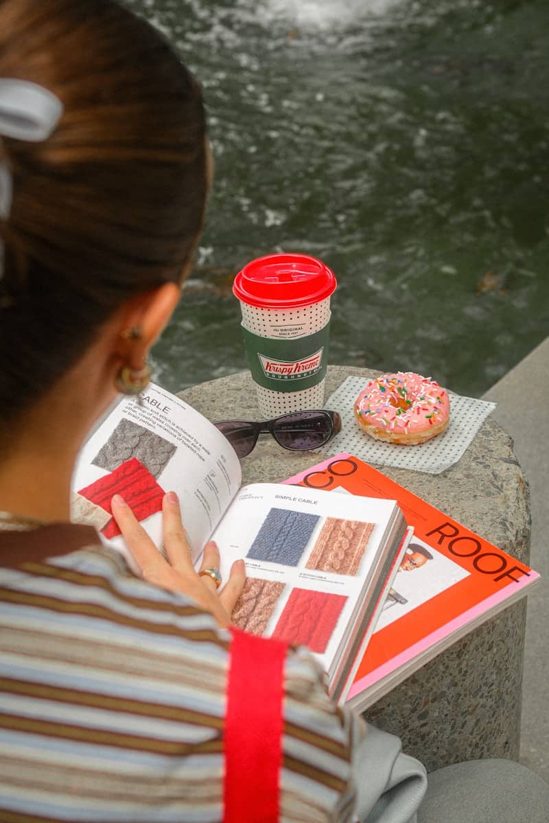 Woman reading a book beside coffee and a pink donut.