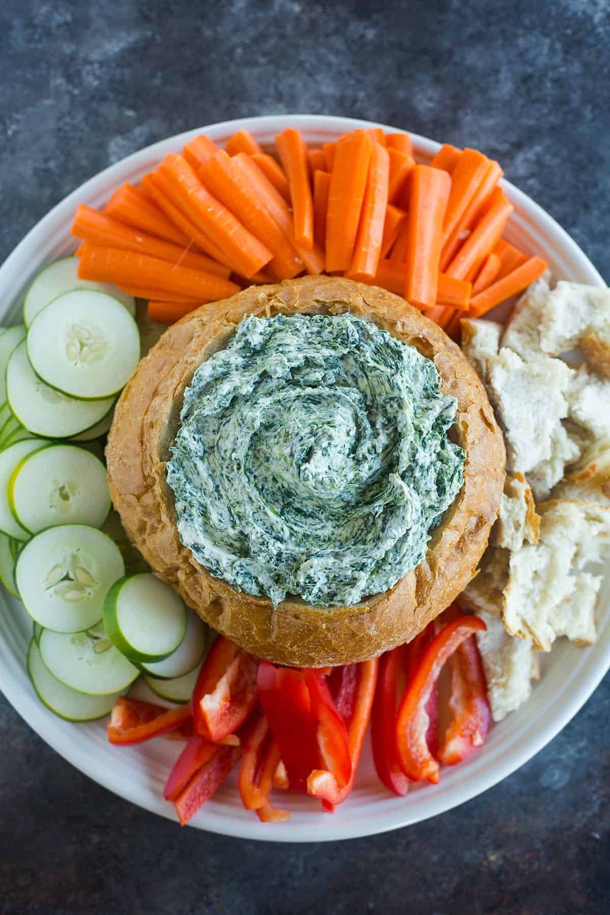 Overhead image of spinach dip in a bread bowl on a platter surrounded by veggies and pieces of bread.