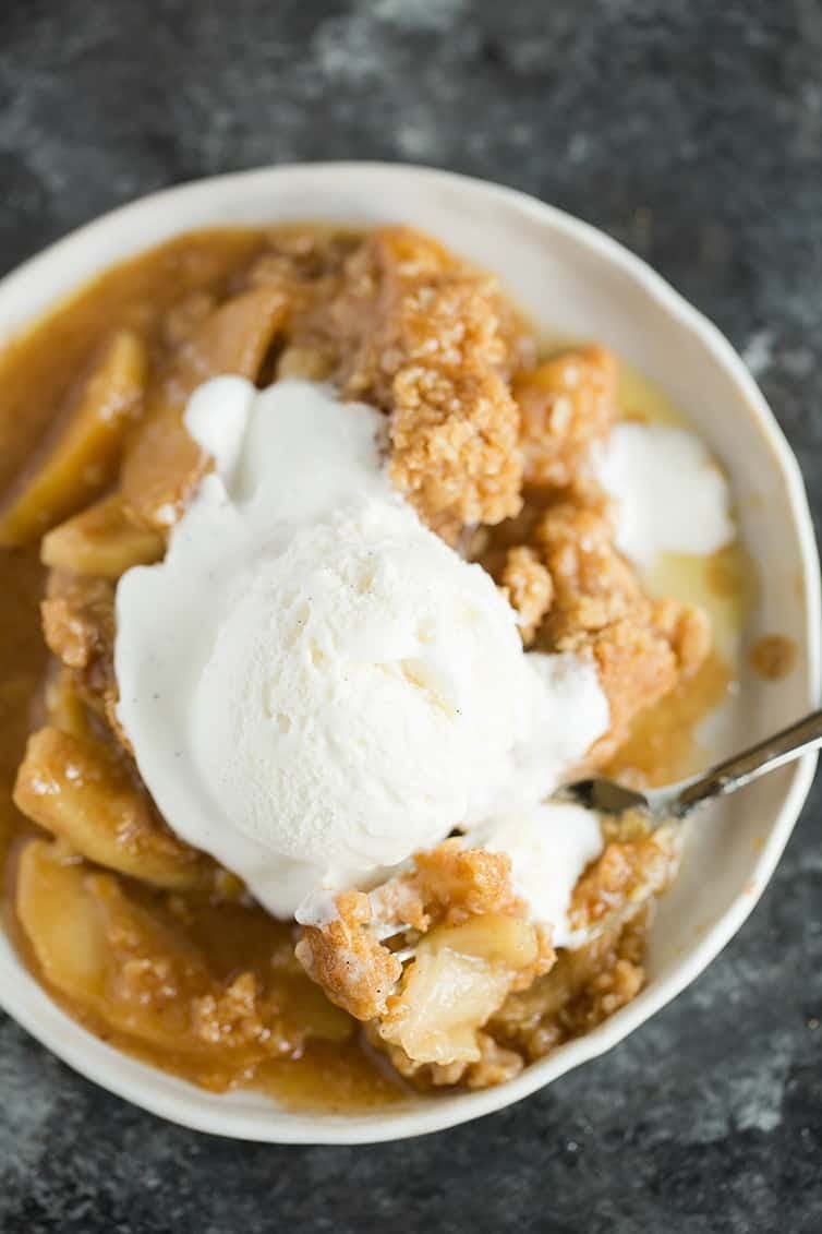 Overhead image of a serving of apple crisp topped with a scoop of vanilla ice cream in a white bowl with a spoon.