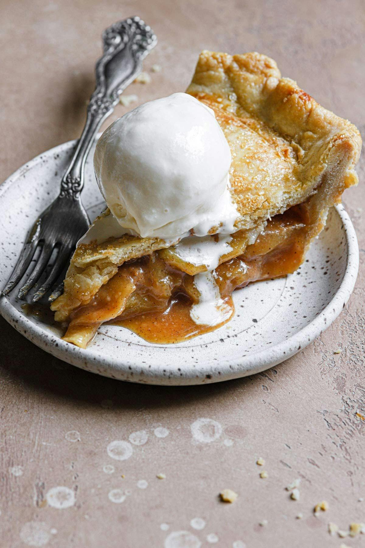 Slice of salted caramel apple pie topped with a scoop of vanilla ice cream on a white plate with a fork.