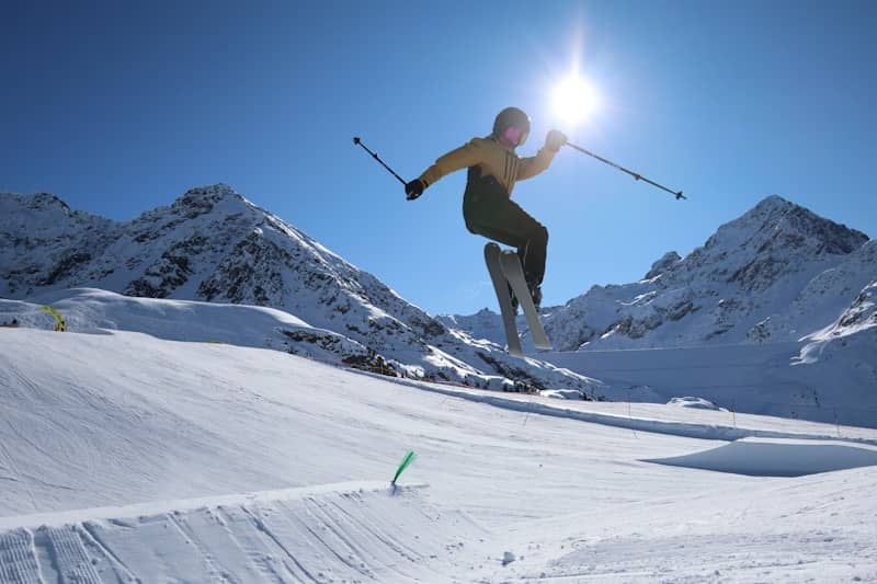 A skier jumping over a snow ramp against a bright sun and mountains