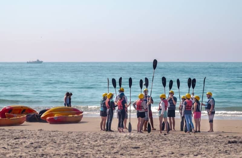 Kayakers gather on a sandy beach with paddles and boats.