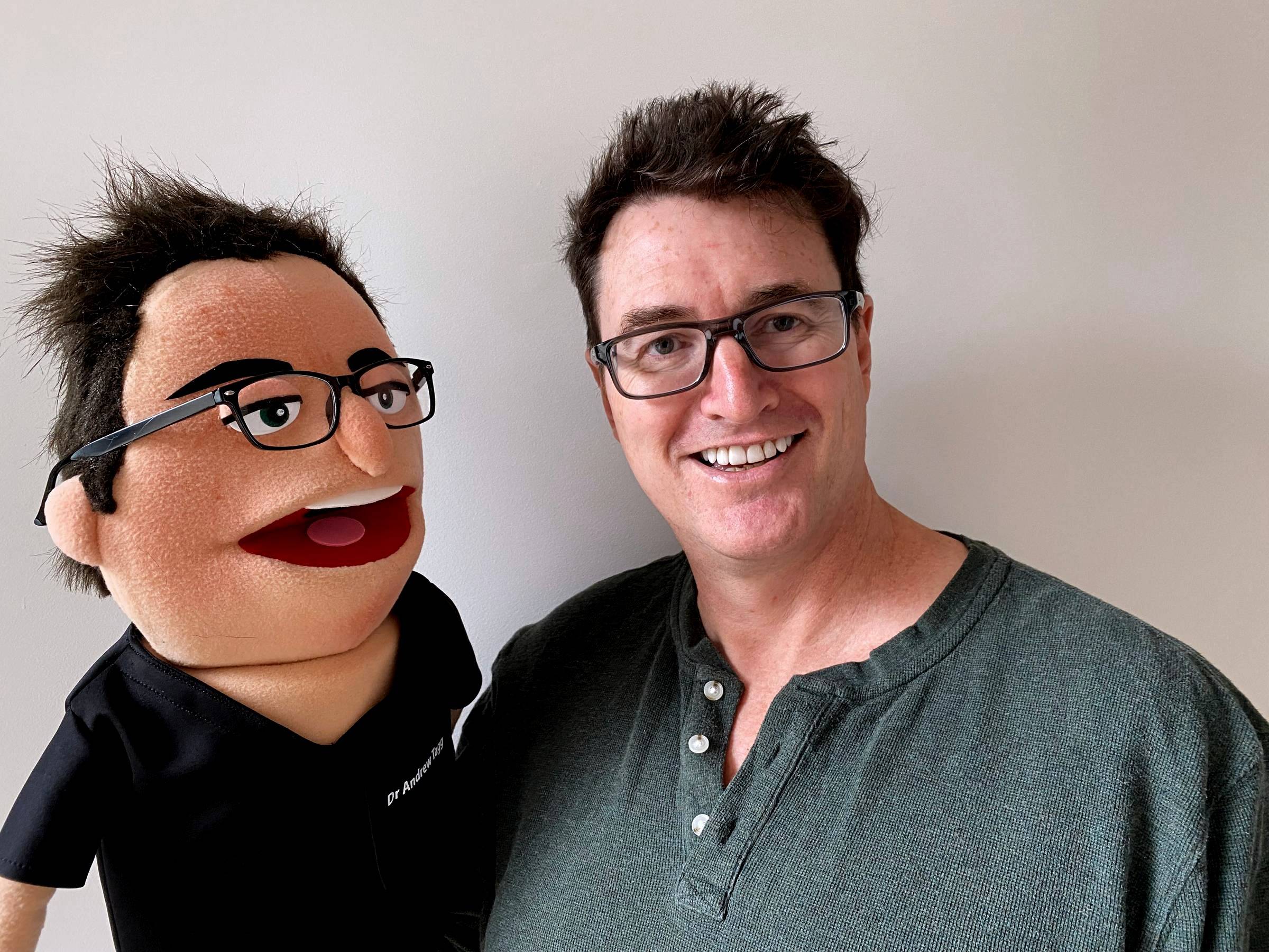 Andy smiling beside a puppet version of himself with spiky dark hair, black glasses and black scrubs.