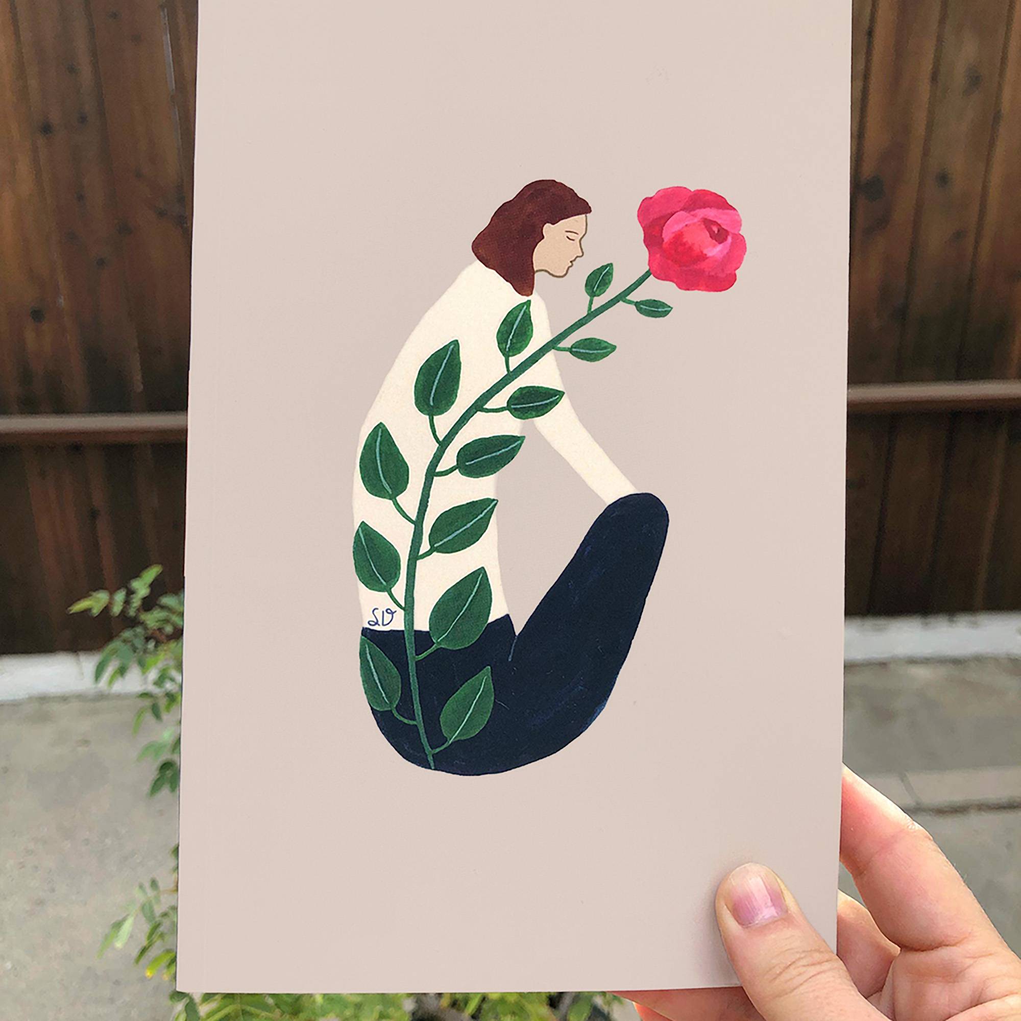 Hand holding journal with illustration of woman profile with eyes closed and rose growing on her spine