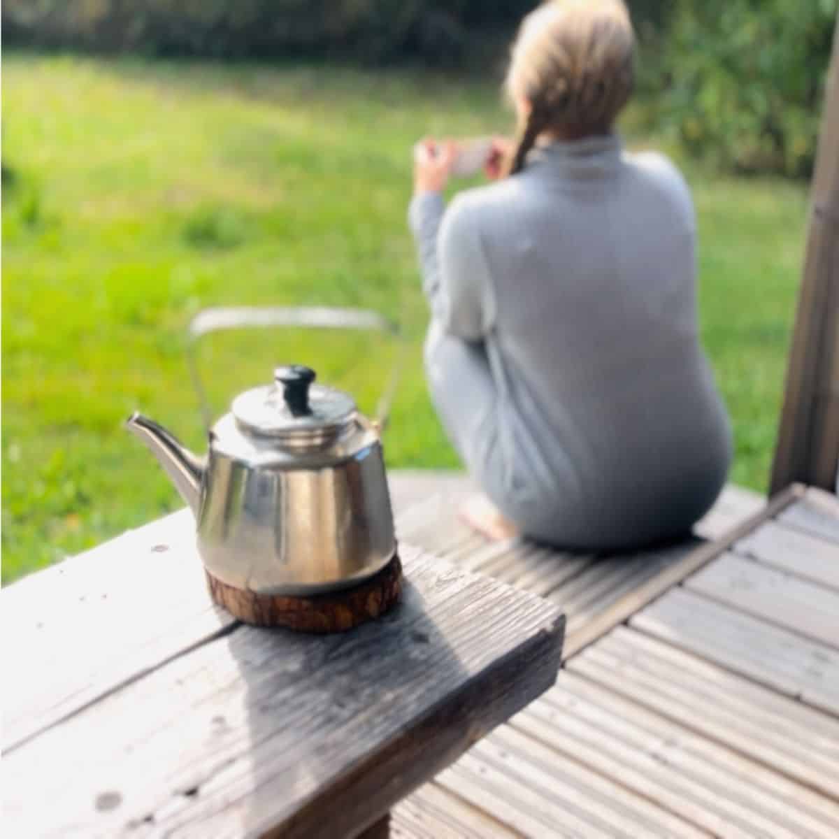 A woman sitting on a porch with a teacup and the tea kettle in on the table.