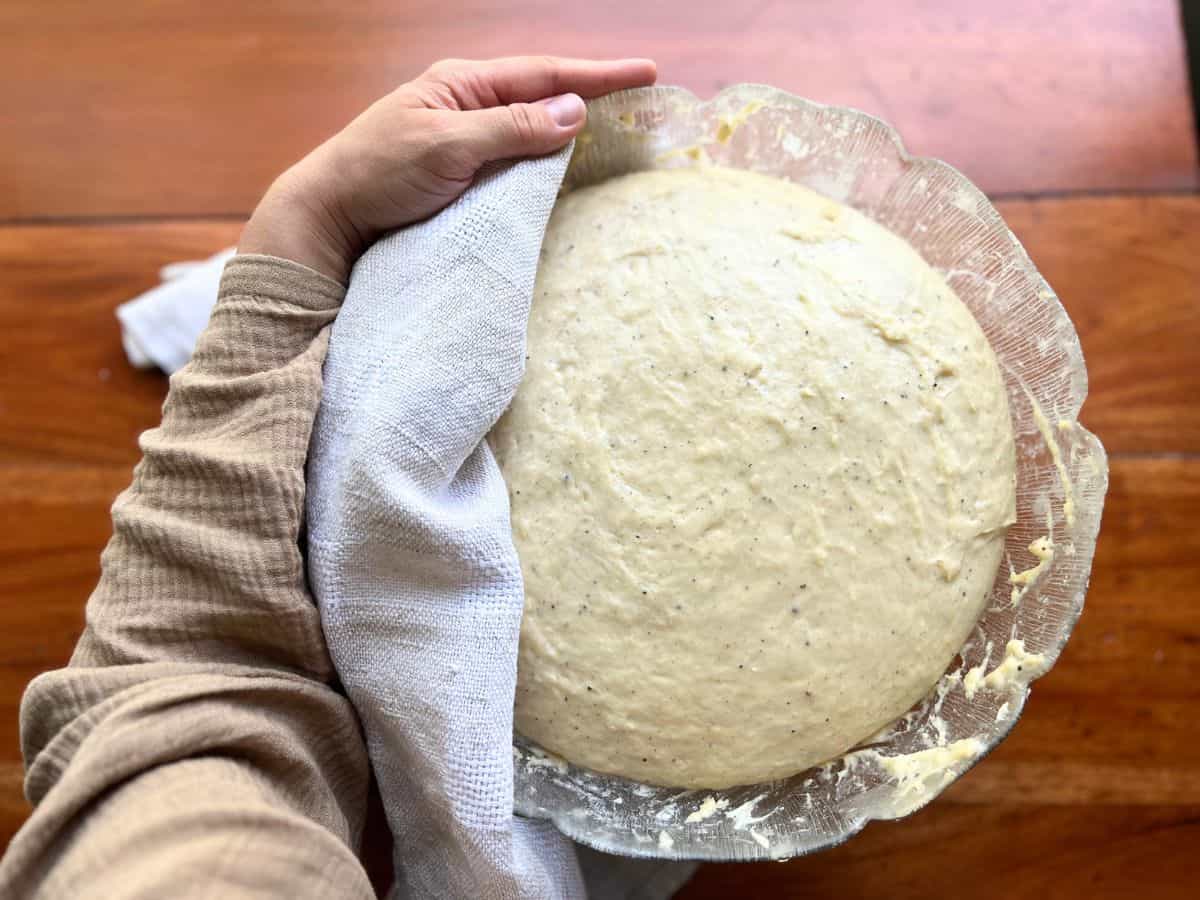 A Nordic sweet bun dough in a bowl and a woman holds it with the tea towel on the side to reveal it.
