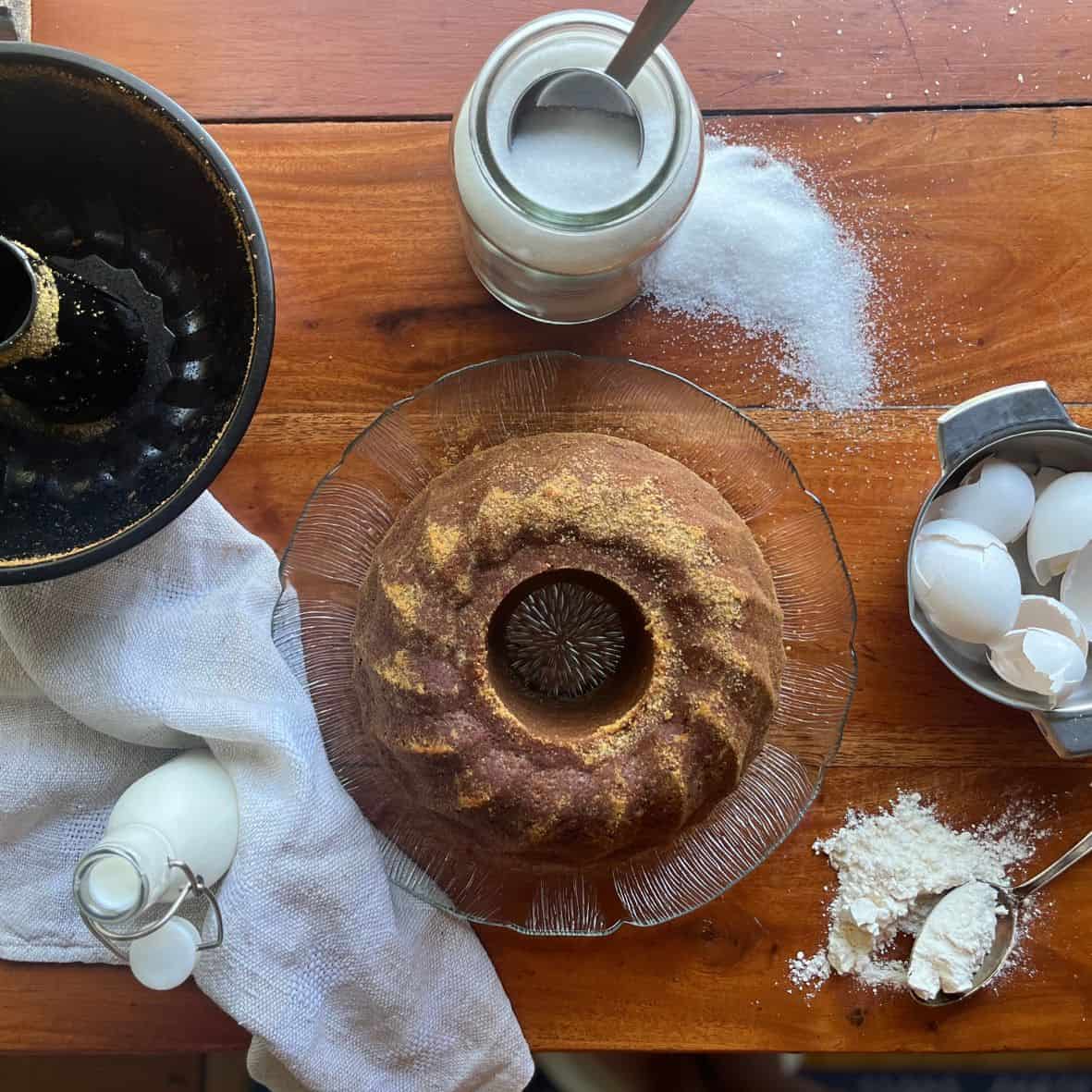 A spiced bundt cake made with kefir on a glass plate with som ecracked egg shells and a jar of sugar on the side.