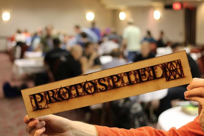 Protospiel sign in the foreground with people gathered around tables at a live event in the background