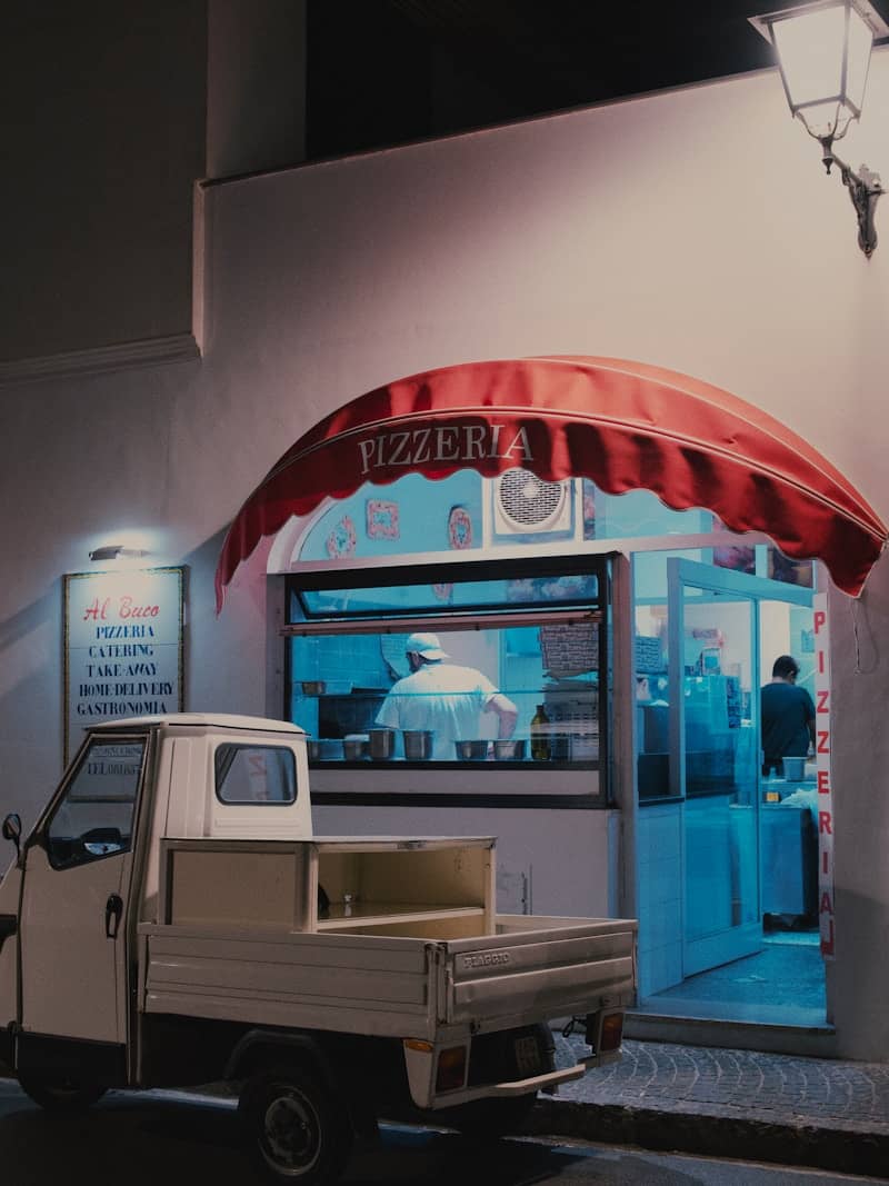 A small pizzeria with a truck at night.