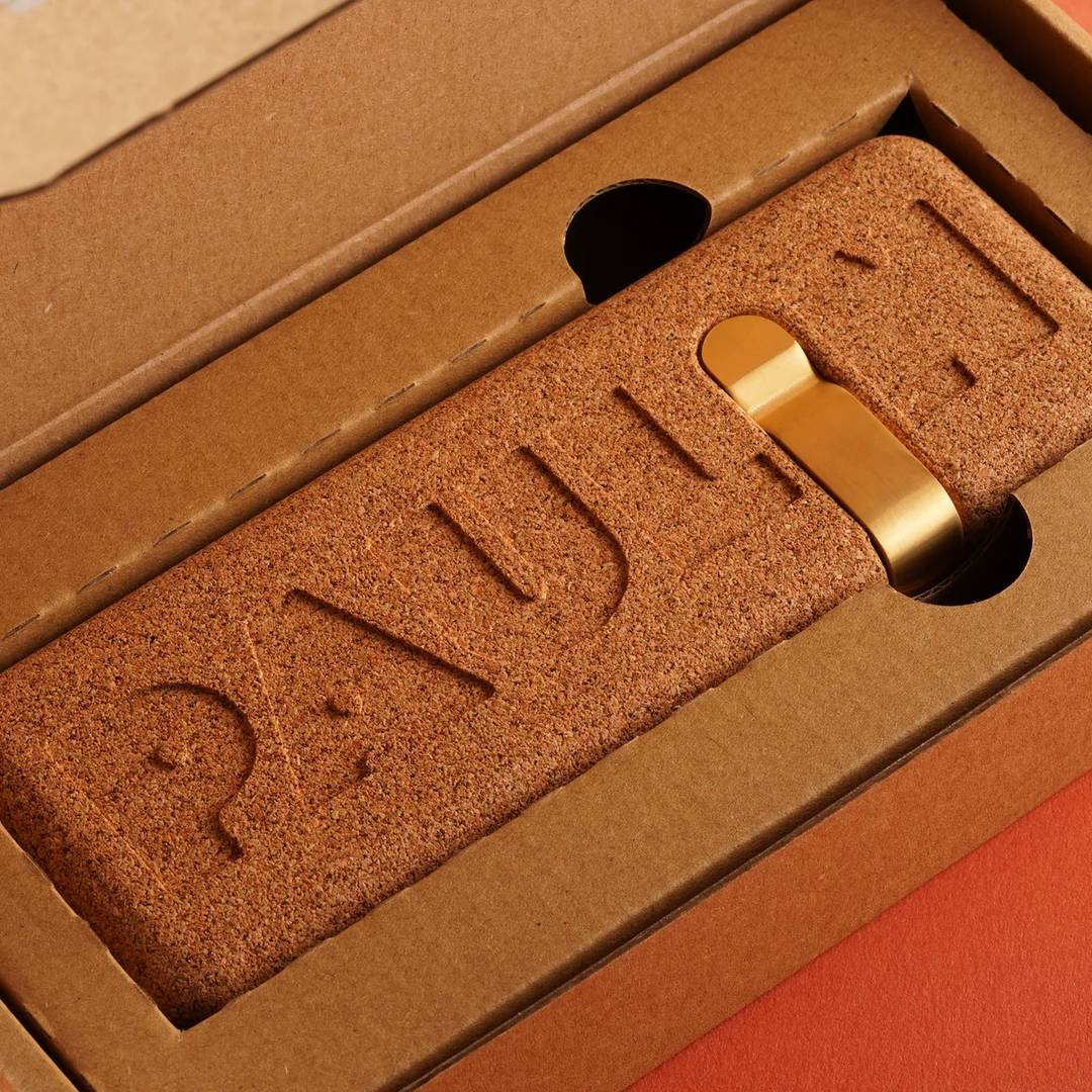 Unboxing view of a Paulin clock showing the brand's thoughtful packaging design. A cork clock back with the Paulin logotype debossed into the material sits in a kraft cardboard tray, with a brass wall-mounting hook visible through a circular cutout. The packaging rests on a coral-colored surface.