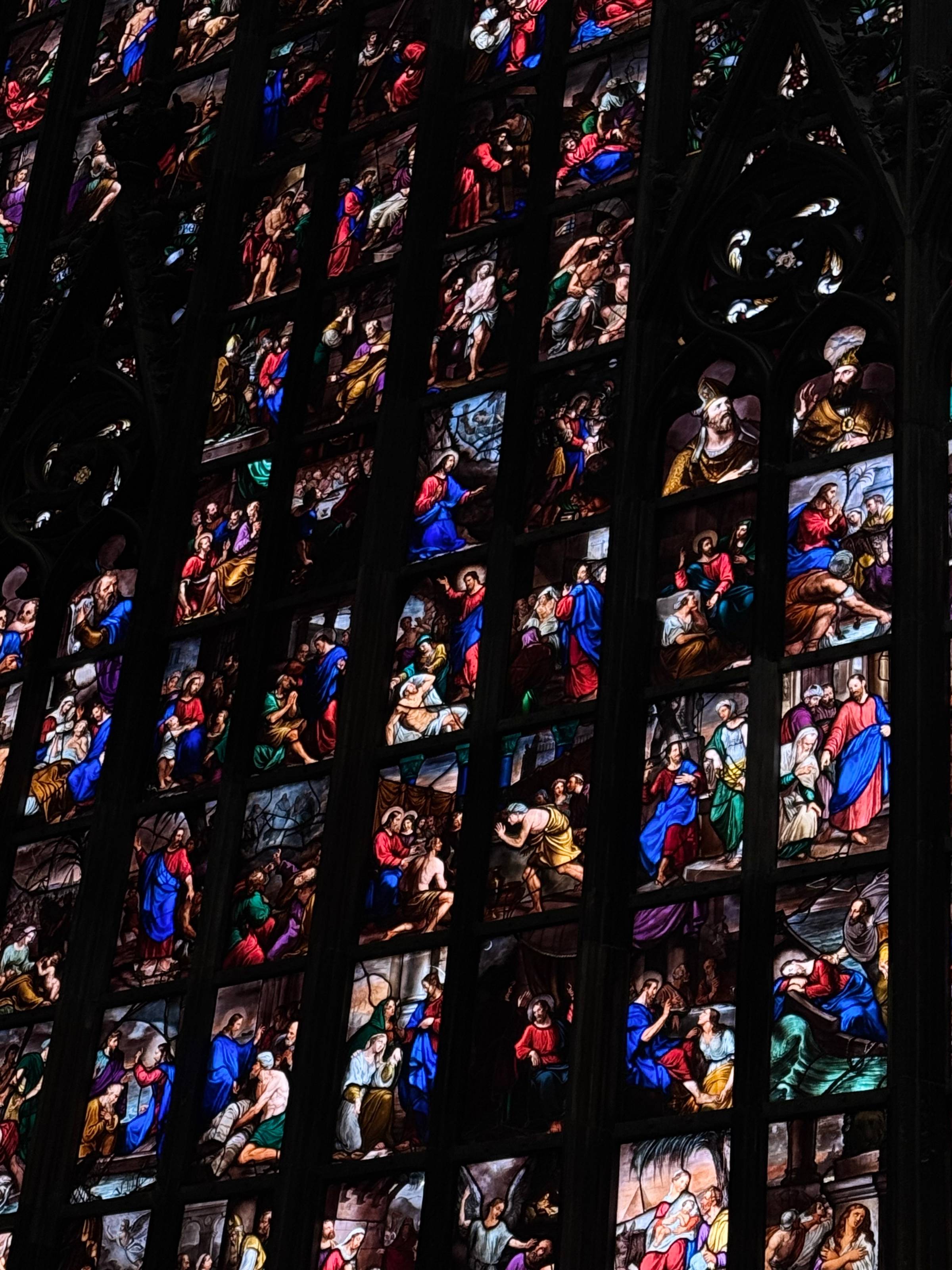 Close-up of towering stained glass windows inside the Milan Duomo, filled with dozens of richly colored biblical narrative scenes in deep blues, reds, and golds, framed by Gothic stone tracery.