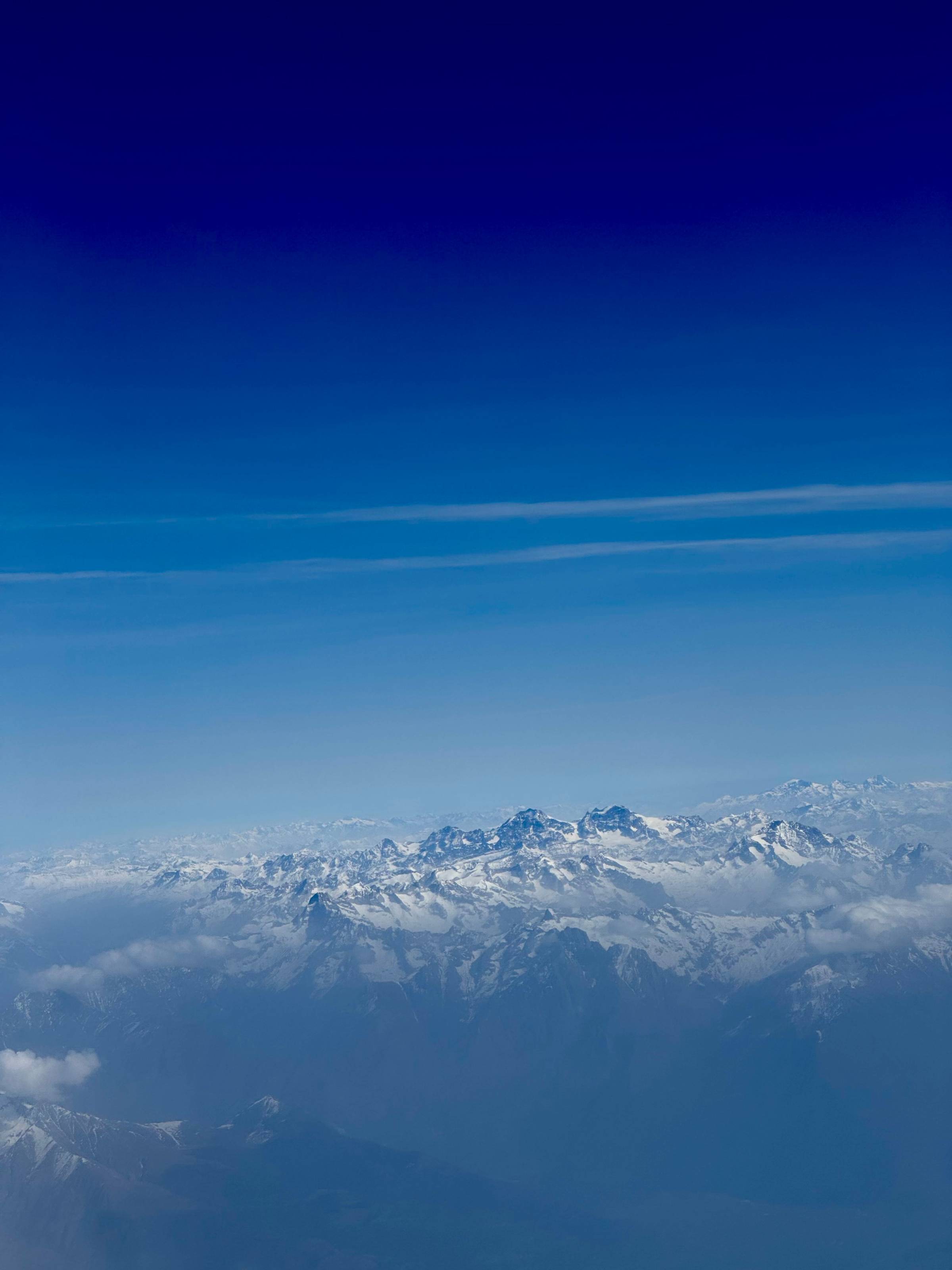 Aerial view of the snow-covered Alps from a plane window, peaks rising above cloud cover into a deep, gradient blue sky.