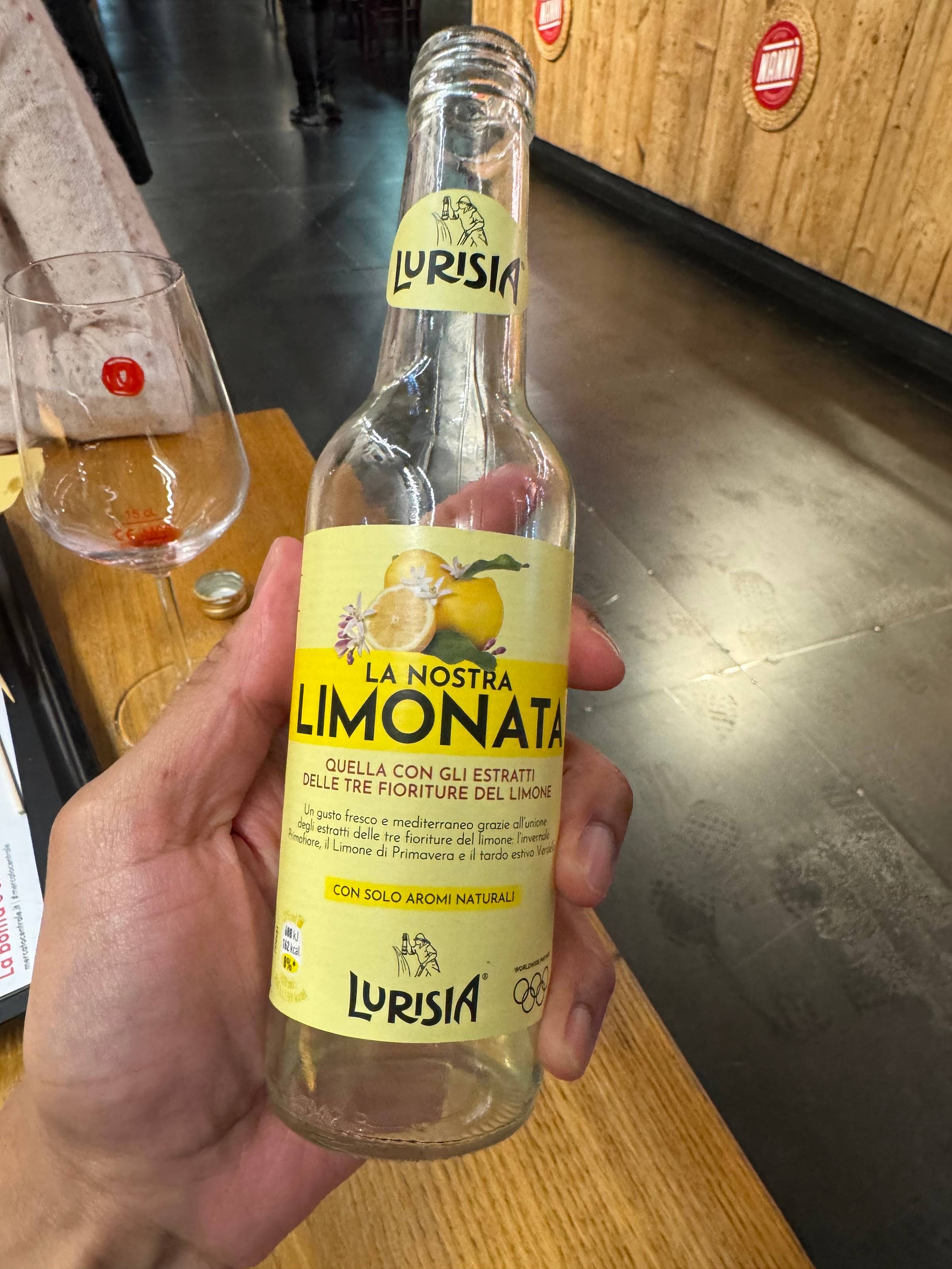 Empty glass bottle of Lurisia La Nostra Limonata, an Italian sparkling lemonade made with extracts from three lemon blossoms, held up against the warm interior of a bar or restaurant.